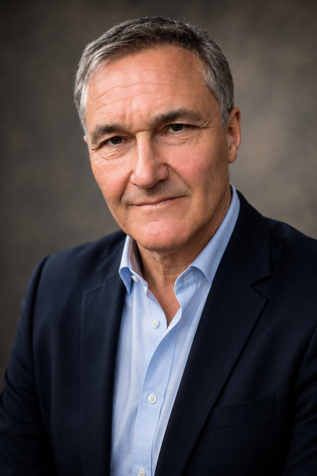 A professional middle-aged man with gray hair, wearing a dark blazer over a light blue collared shirt, looking directly at the camera with a slight smile.