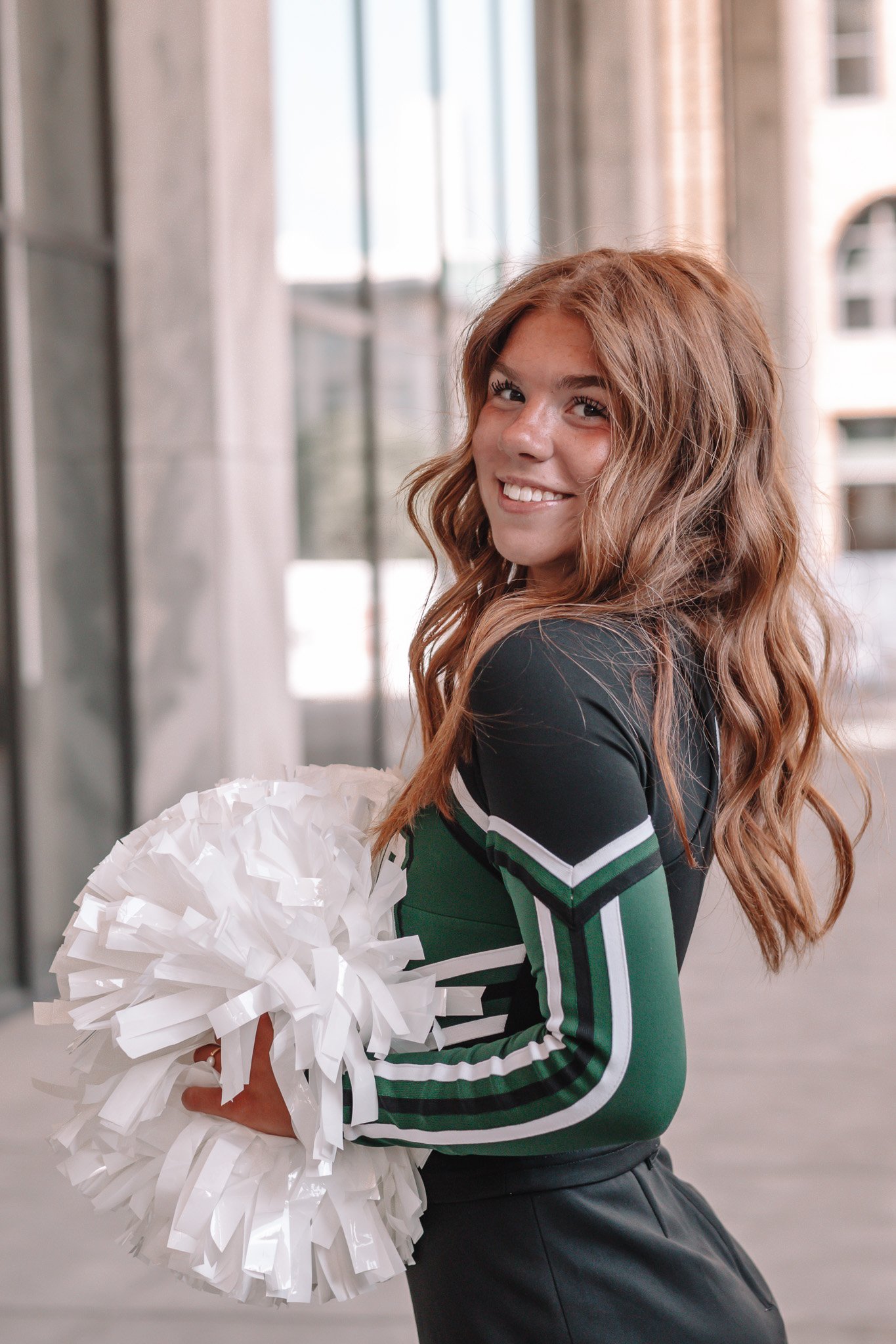 Young woman with long, wavy, reddish-brown hair smiling and holding white pom-poms, wearing a black, green, and white cheerleading uniform, standing indoors near large windows.