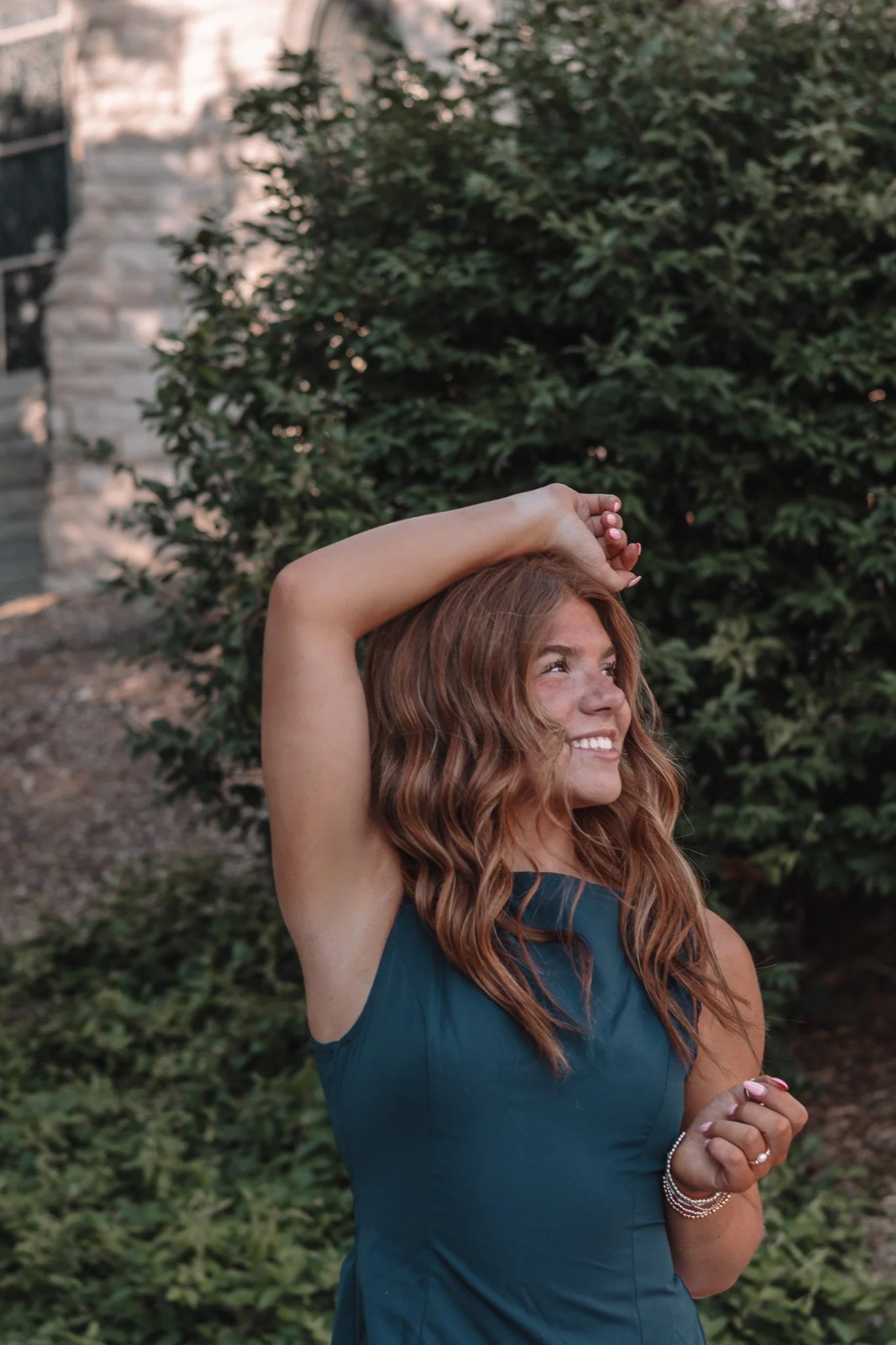 A woman with long, wavy red hair and light skin is smiling outdoors, wearing a sleeveless dark teal dress and jewelry on her wrist, with her right arm resting above her head in front of green foliage.