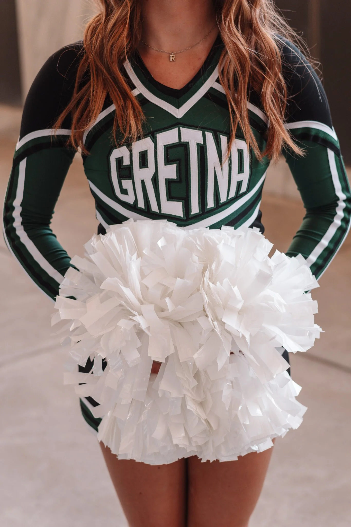 Close-up of a dancer wearing a green and black cheerleading uniform with 'GRETNA' written on it, holding white pom-poms, with legs and part of a blurry background visible.