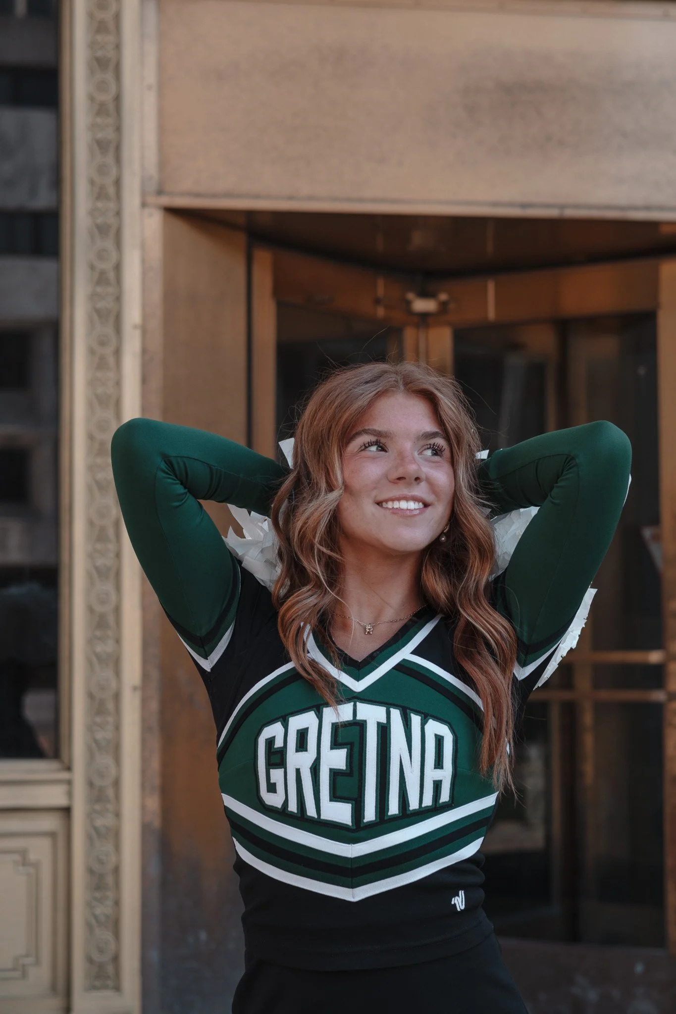 Young woman smiling, wearing a green and black cheerleading uniform with 'Grefina' on it, standing outside in front of a building.