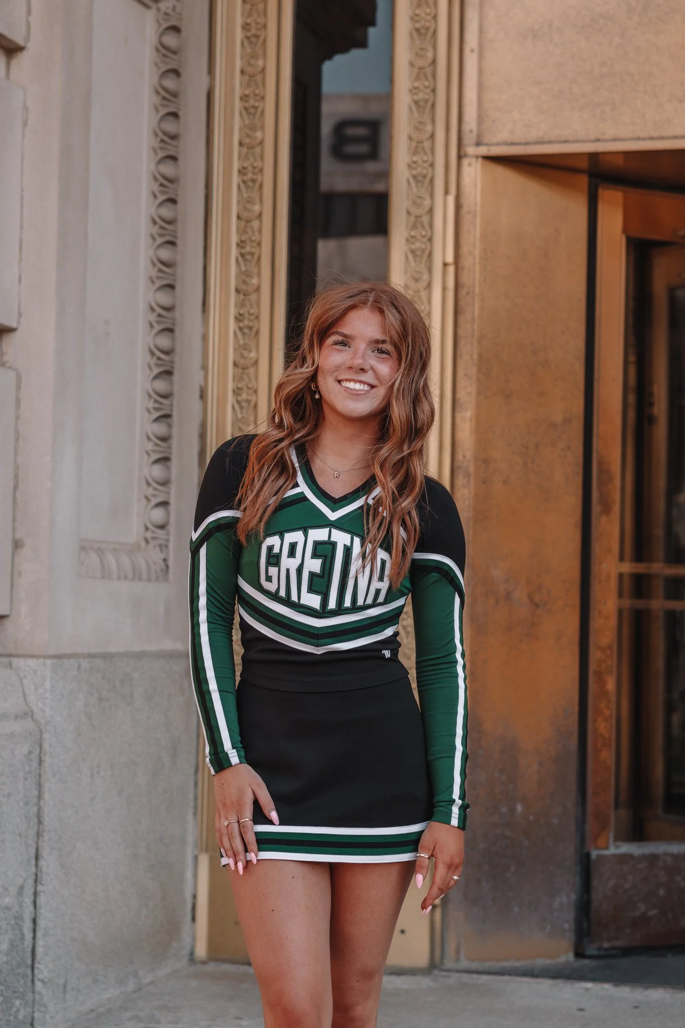 A young woman with long wavy auburn hair smiling and wearing a green and black cheerleading uniform with the word "GREAT" on it. She is standing outside near a building with ornate architectural detailing. The woman is showing her legs and has rings 