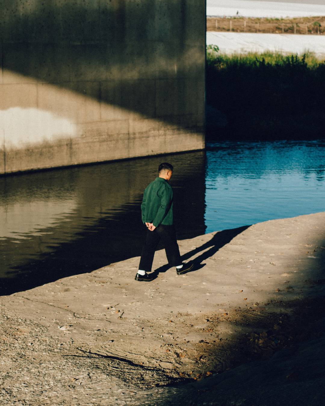 Photo of Chris Datiles, Los Angeles IFS Therapist, wearing a dark green jacket, black trousers, white socks, and dark shoes walking along a concrete ledge beside the LA River. His back is to the camera, hands clasped behind him.