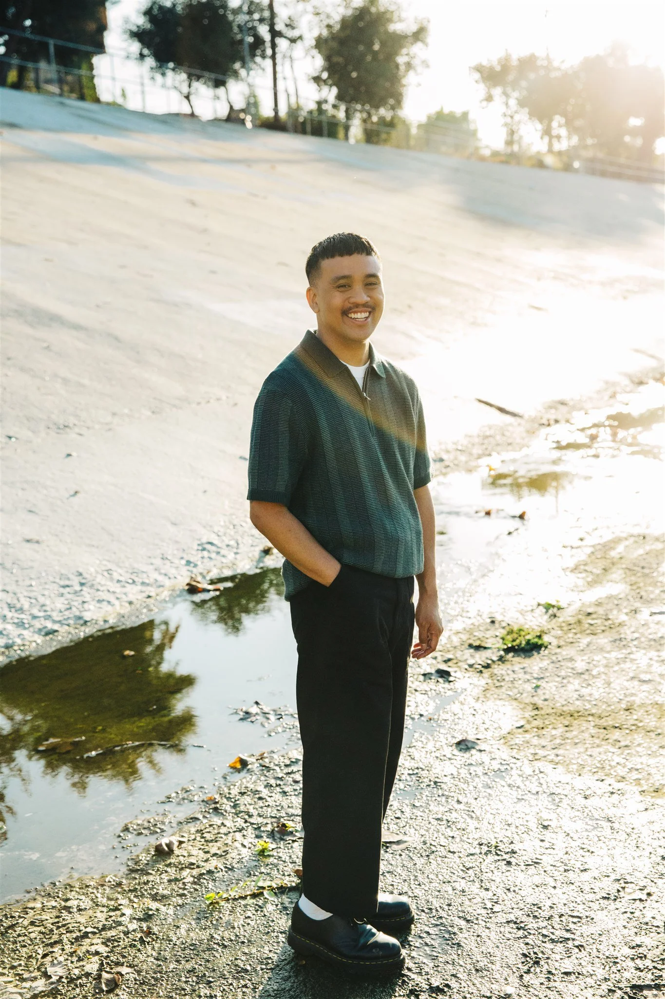 Portrait of Chris Datiles, LGBTQ Los Angeles IFS Therapist, standing on concrete near the LA river. Chris is a brown Filipino American, smiling at the LA river with the sun setting.