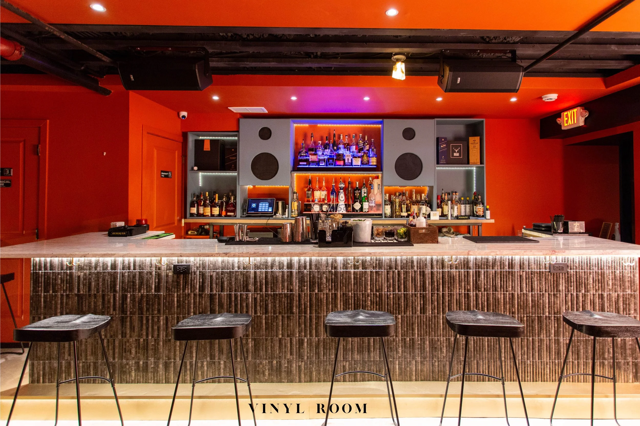 Bar with a marble countertop, wooden paneling, and four black bar stools. Shelves behind the bar display bottles of alcohol, with some illuminated by blue and orange lights. Orange-painted walls and ceiling with recessed lighting and speakers.