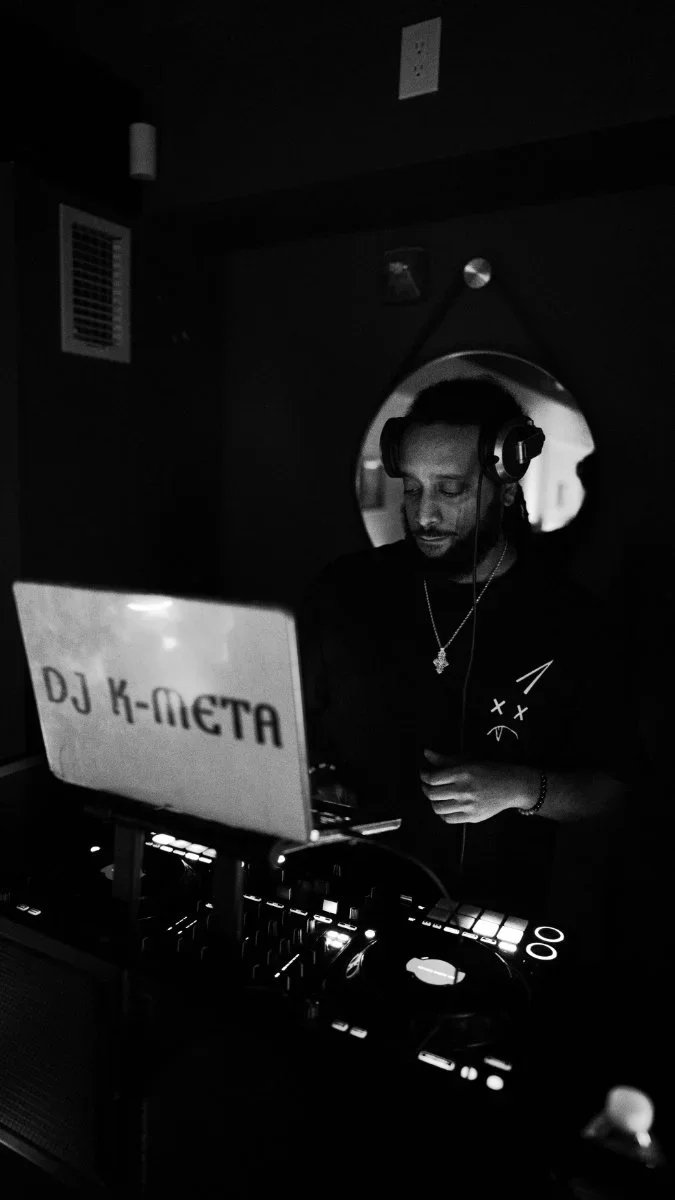 A DJ wearing headphones is at a mixing console, with a circular mirror reflecting his face, in a dark room with a sign that reads 'DJ K-META'.
