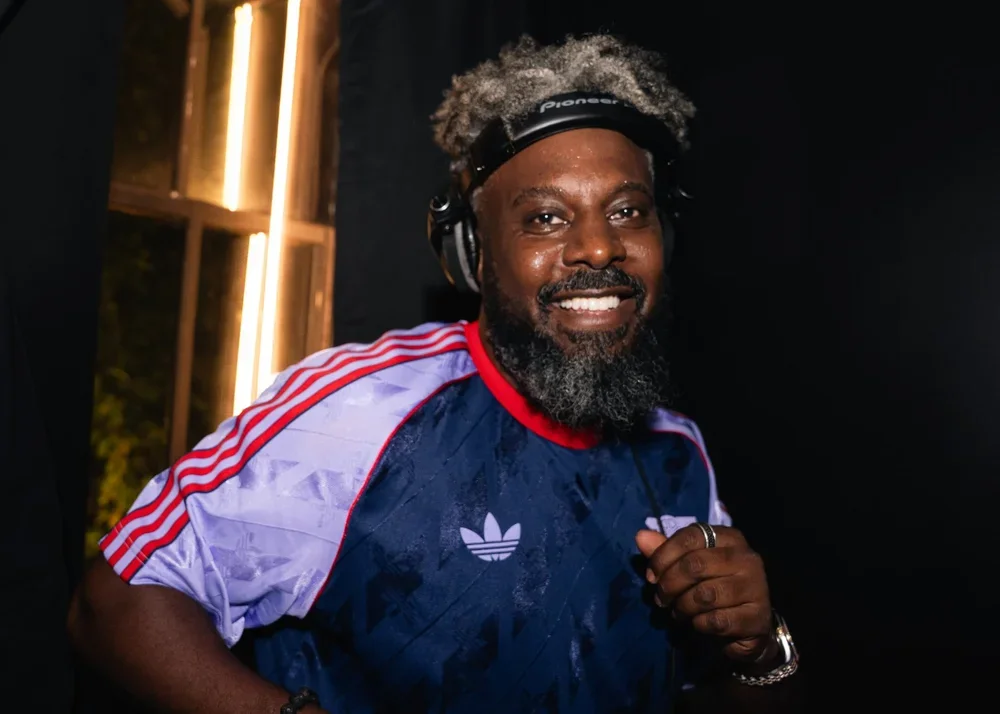 A smiling man with gray hair and a beard, wearing a black Pioneer headphones, a blue Adidas sports jersey with red and white stripes, and a ring on his right hand, standing indoors with a black background and a wooden frame with vertical lights behind him.
