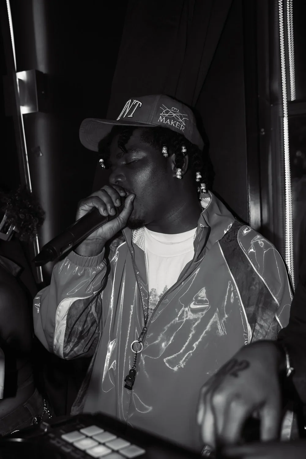 A young man with dark skin, braided hair with beads, is singing or rapping into a microphone. He is wearing a cap and a shiny, reflective jacket. The background is dark and appears to be indoors.