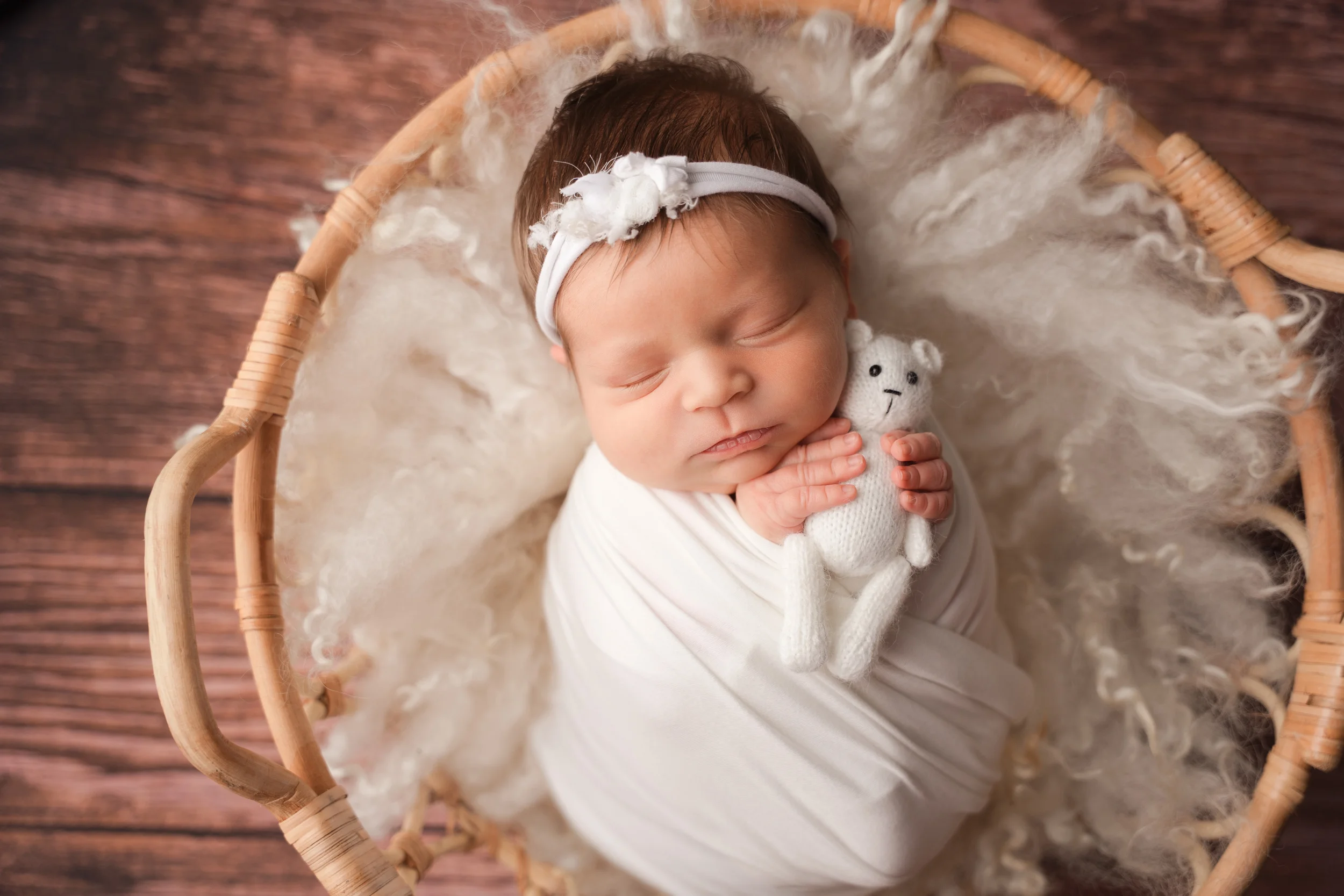 newborn baby girl wrapped in white with small white stuffed bear posed in a basket with a white layer during in home session with raleigh newborn photographer