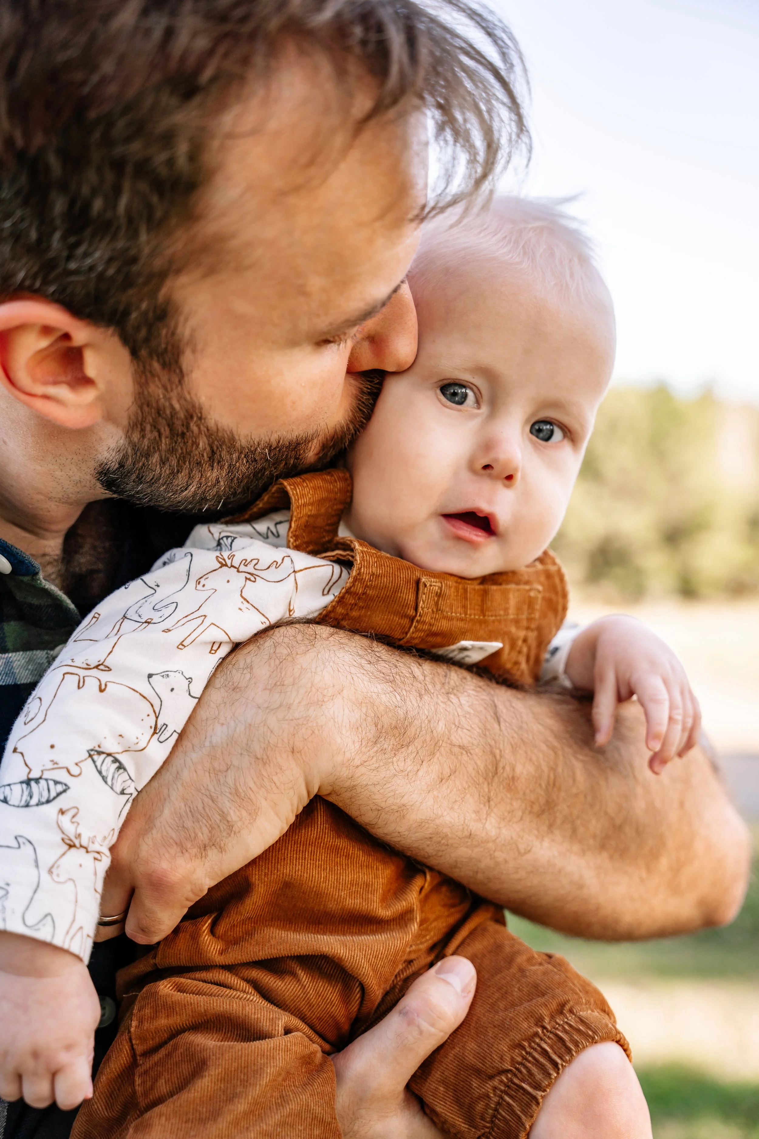 A man holding a young child outdoors, the man is kissing the child's cheek, and the child is looking at the camera.