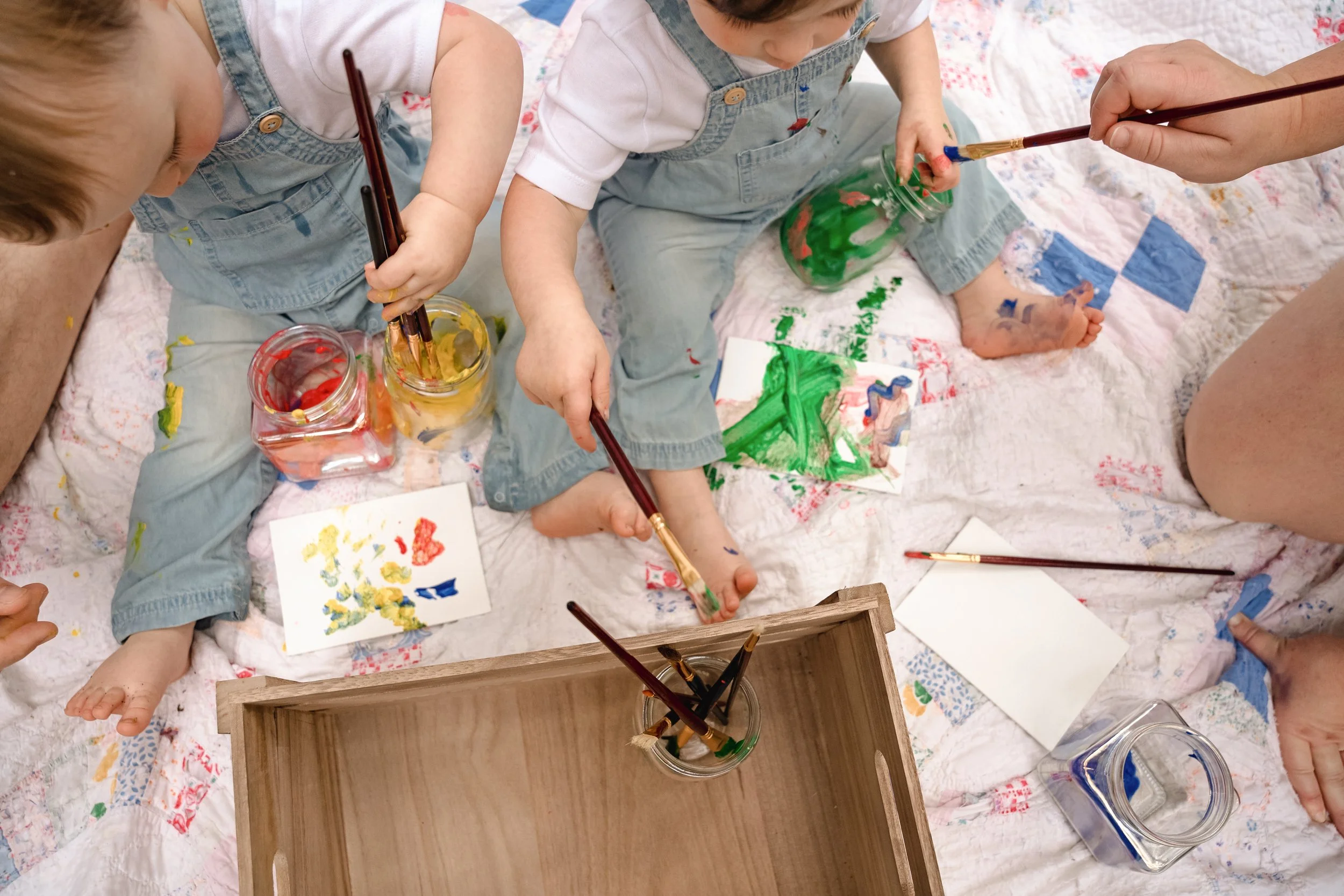 Two young children painting with brushes in jars of water, seated on a quilt with open watercolor paints and paper around them.