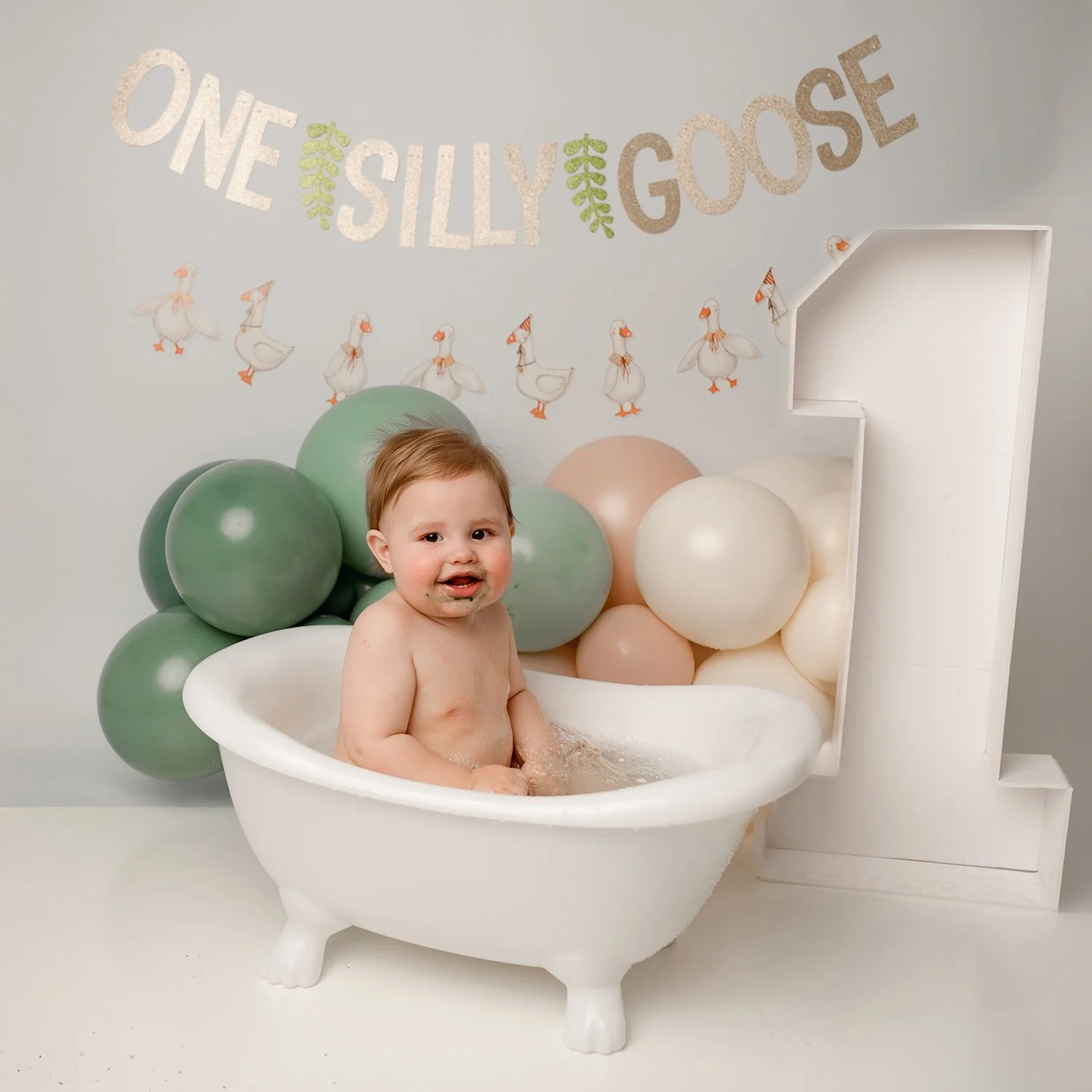 Baby sitting in a small white bathtub with balloons and a backdrop saying 'One Silly Goose' with duck illustrations.