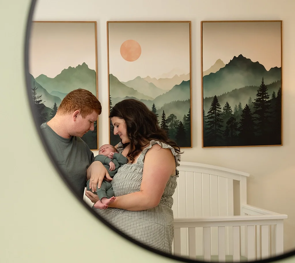 A couple holding a newborn baby in a nursery with mountain and forest wall art during session with cary newborn photographer