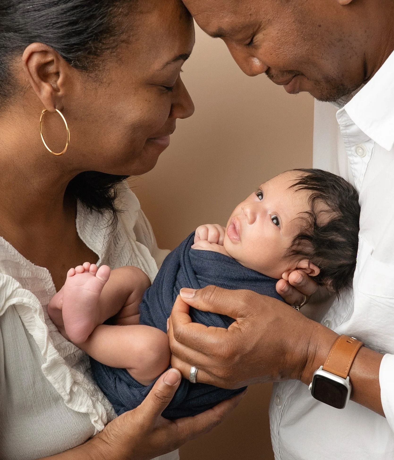 A close-up of a family moment showing an adult woman, an adult man, and a young child. The woman and man are touching foreheads and smiling gently at the child, who is gazing up at them during session with Raleigh Newborn Photographer
