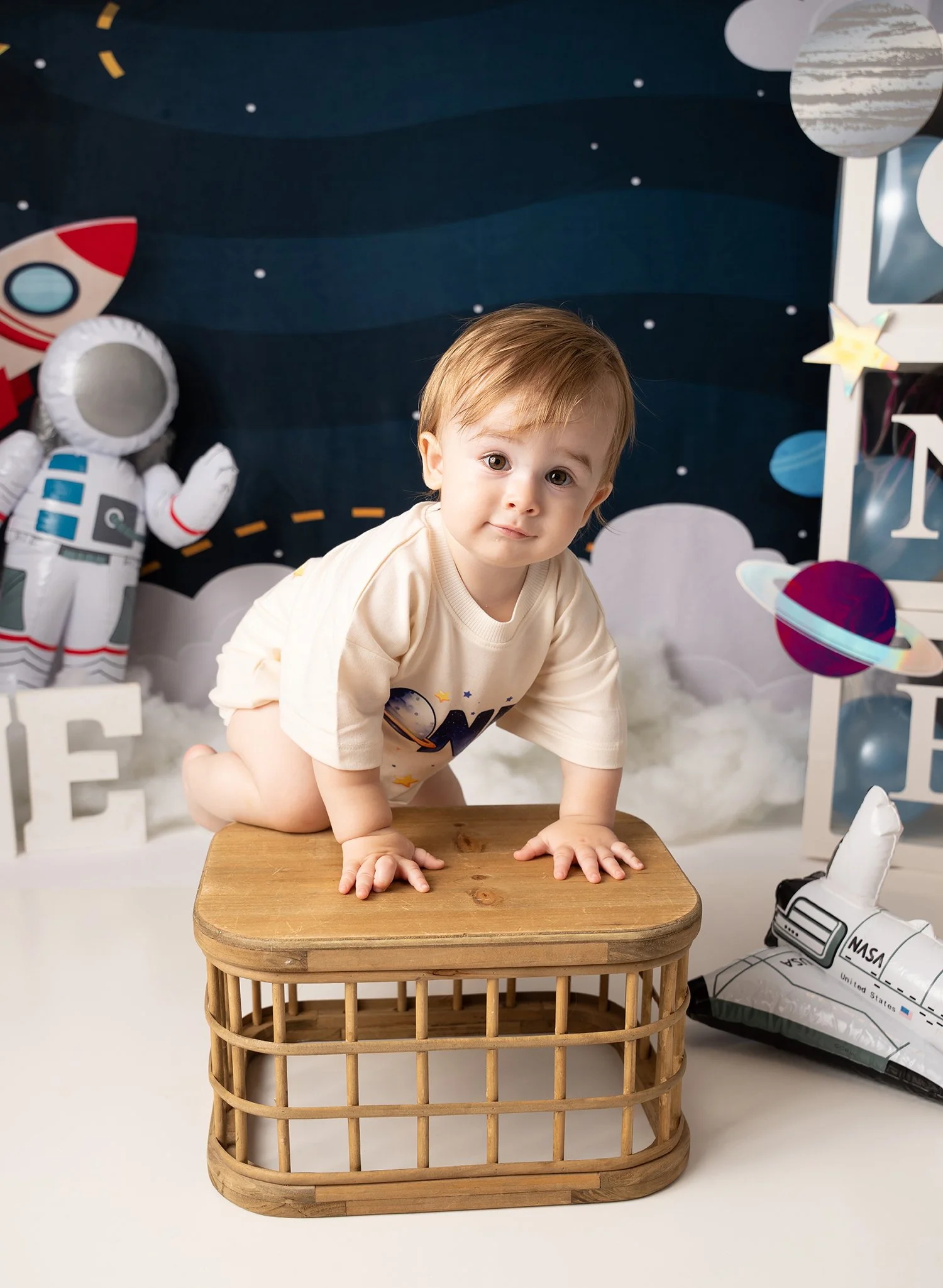 A young child crawling on a wooden side table in space-themed setting with NASA balloon, astronaut plush, and planets decorations.