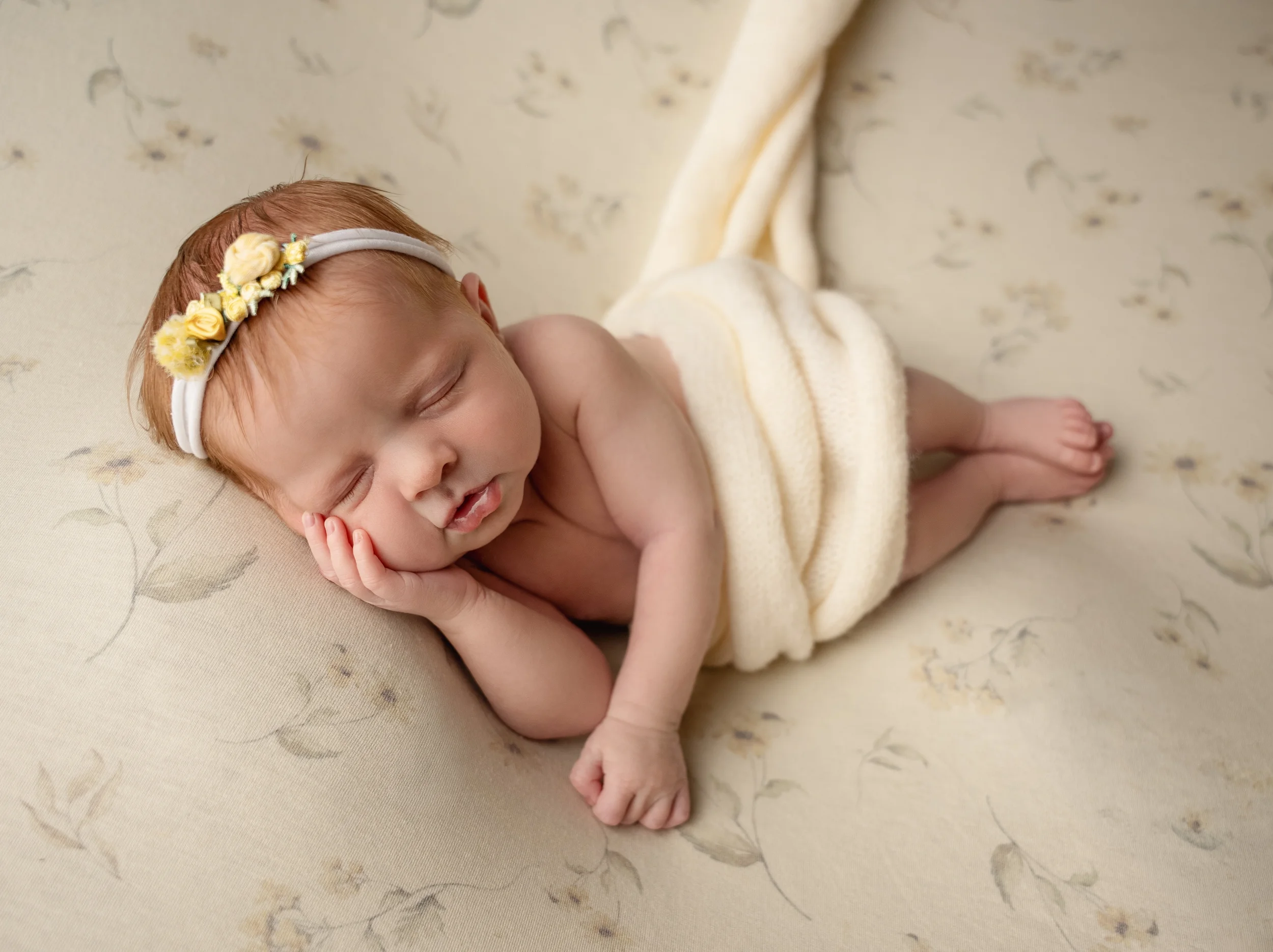 A sleeping baby with red hair, wearing a floral headband and wrapped in a yellow towel, lying on a patterned cream-colored surface during session with Cary newborn Photographer