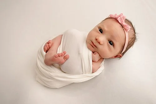 A baby girl wrapped in a white blanket with a pink bow headband, lying on a flat surface during session with apex newborn photographer