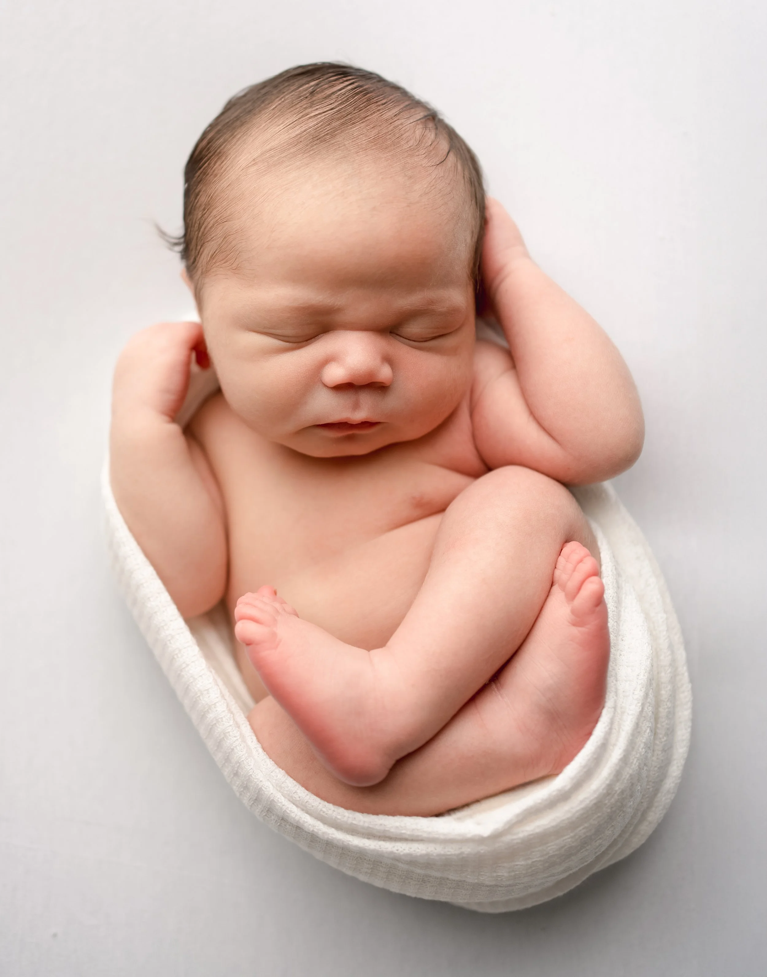 A newborn baby with closed eyes, curled up, and resting while being gently held in hands, wrapped in a white cloth, against a plain white background.