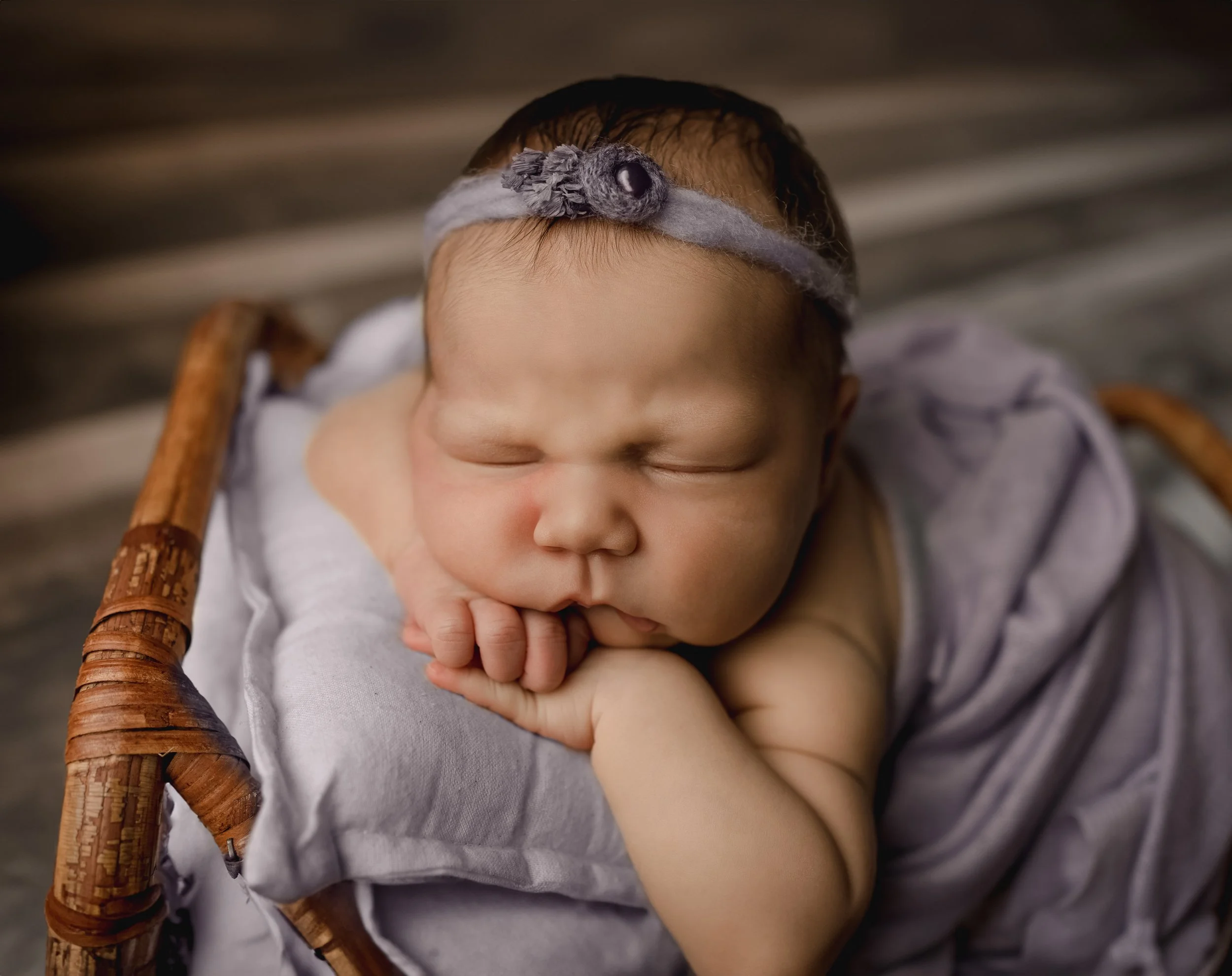 Close-up of a sleeping baby with hands near face, wearing a headband with gray flower, resting on a woven chair.