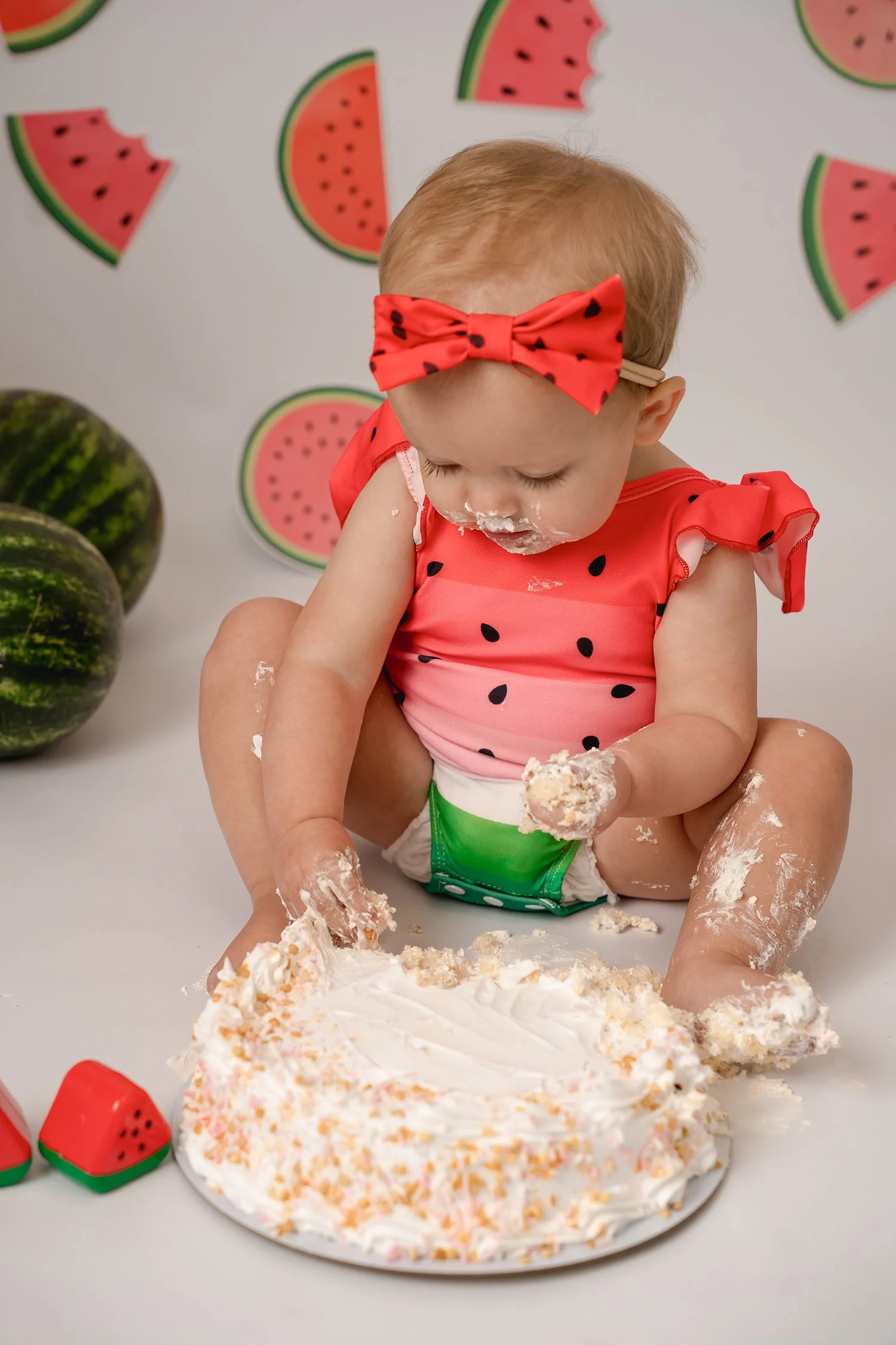 A young child in a watermelon-themed outfit with a red bow headband sat on the floor, playing with a birthday cake covered in white frosting. The background has watermelon slice decorations, and there are real watermelons beside the child.
