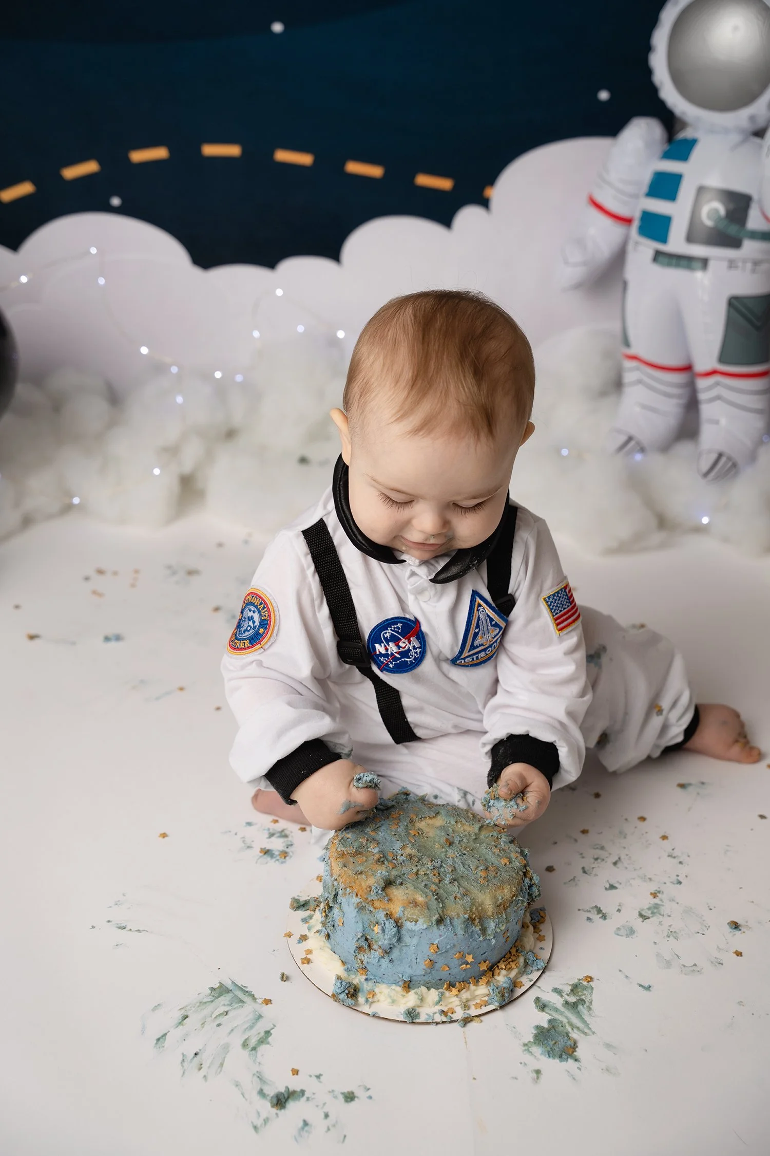 A young child in a space-themed costume sitting on the floor, smashing a blue birthday cake with a NASA logo, with space-themed decorations in the background.
