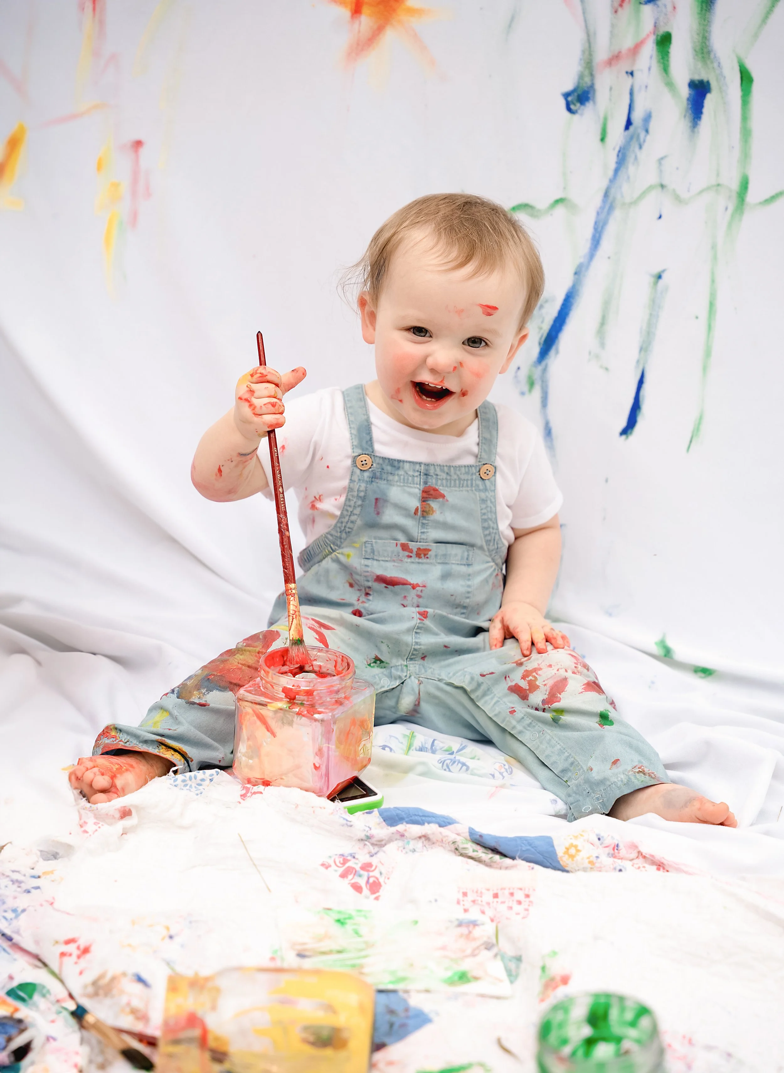 A young boy with light brown hair, wearing overalls and a white shirt, is sitting on a white sheet covered in colorful paint, holding a paintbrush and smiling at the camera with paint on his face, hands, and clothes, surrounded by splattered paint an