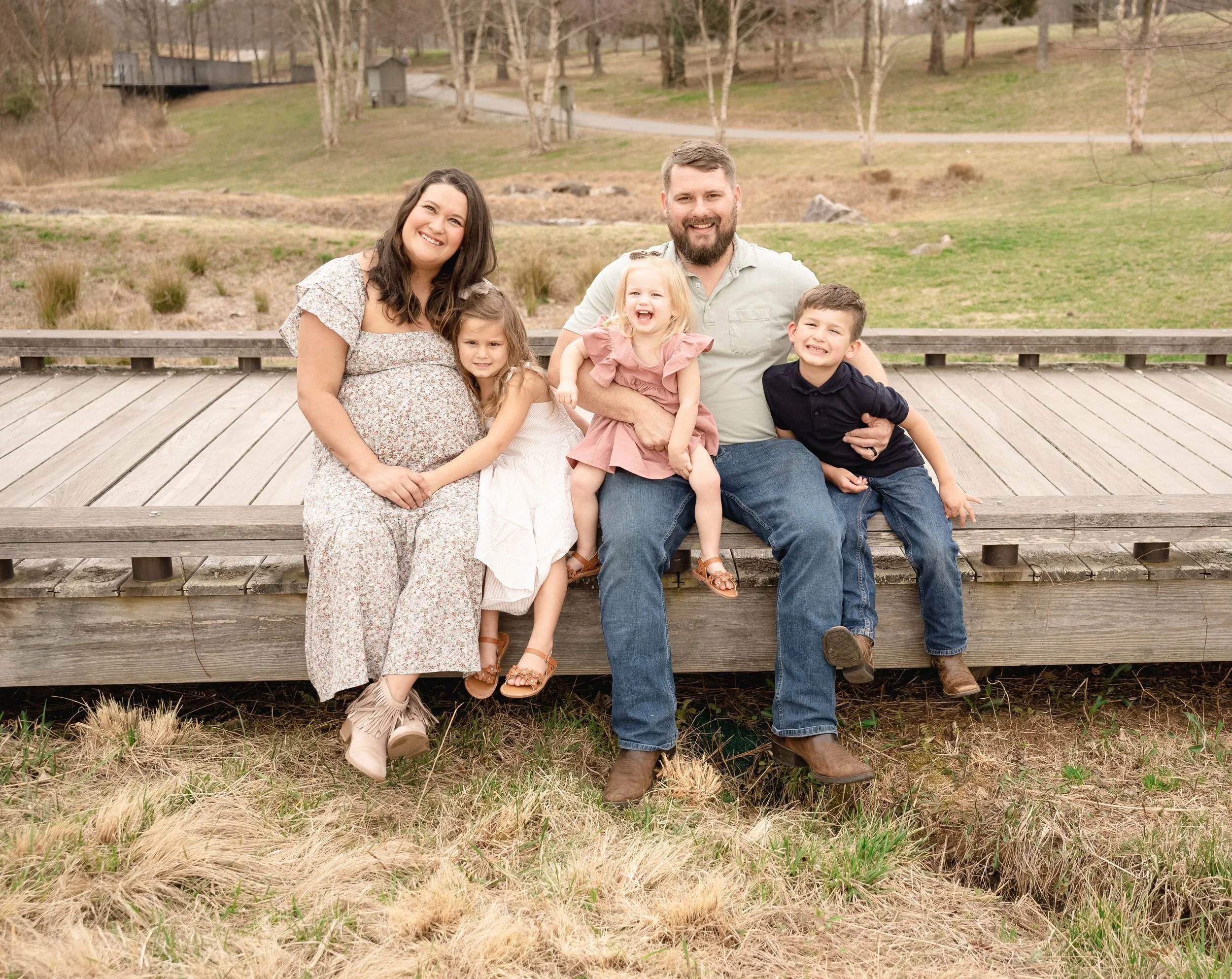 A family of five sitting on a wooden bridge outdoors, smiling, with trees and grass in the background.