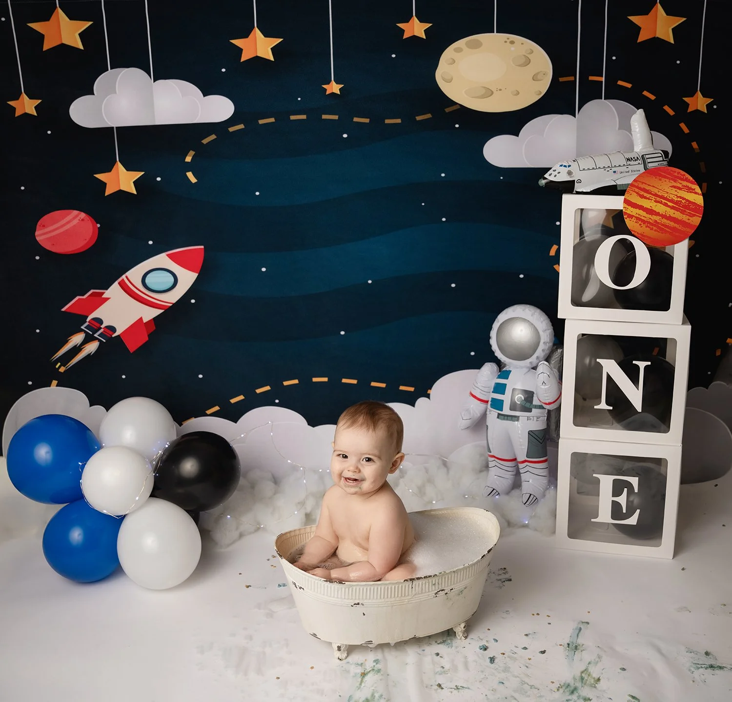 A baby sitting in a bathtub filled with foam, surrounded by space-themed decorations including balloons, stars, an astronaut figure, and a rocket, against a space backdrop with planets and clouds.