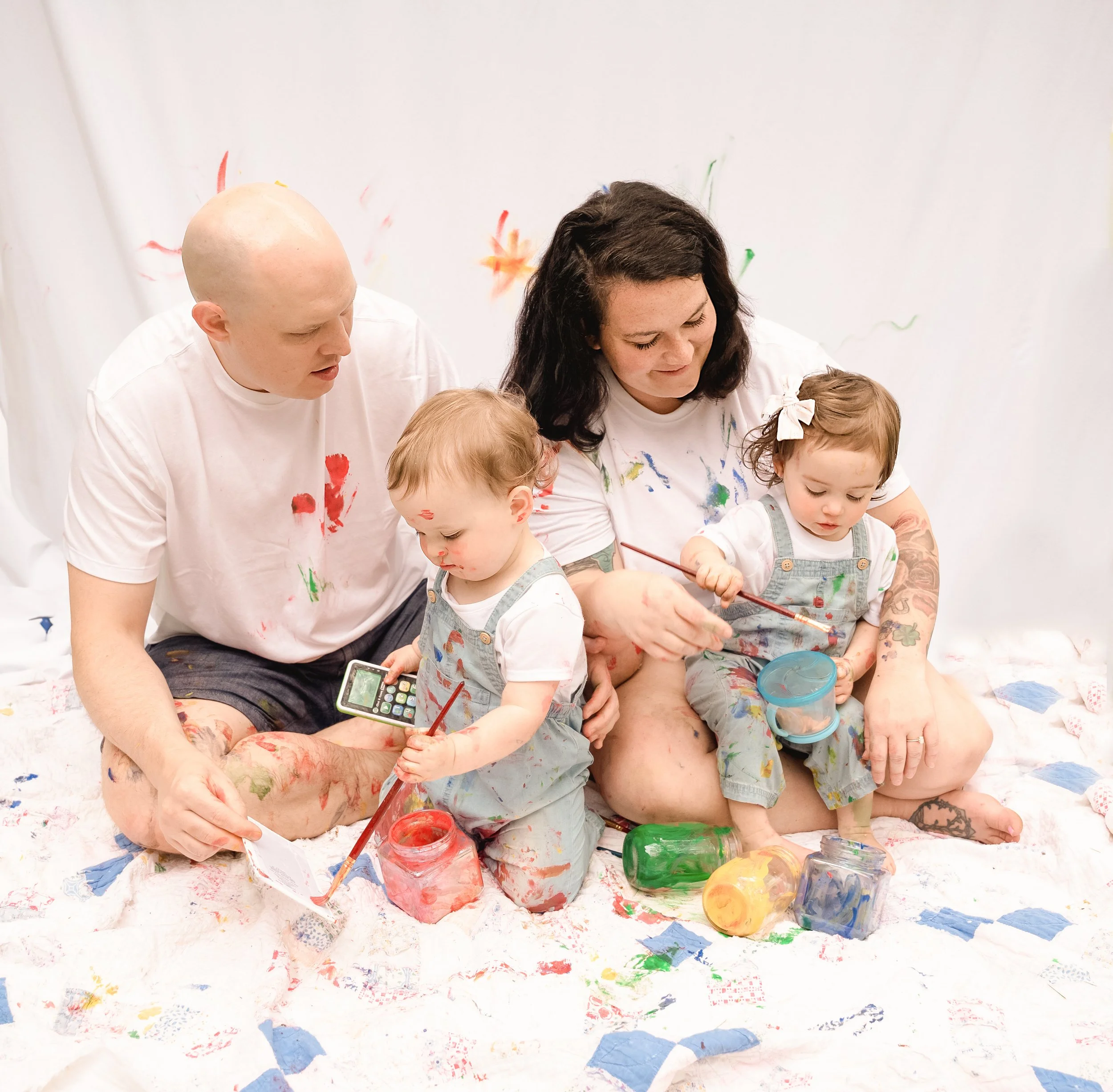 Family of four, two adults and two children, with paint on their skin and clothes, sitting on a white sheet with colorful paint jars, painting on a white background during session with raleigh family photographer