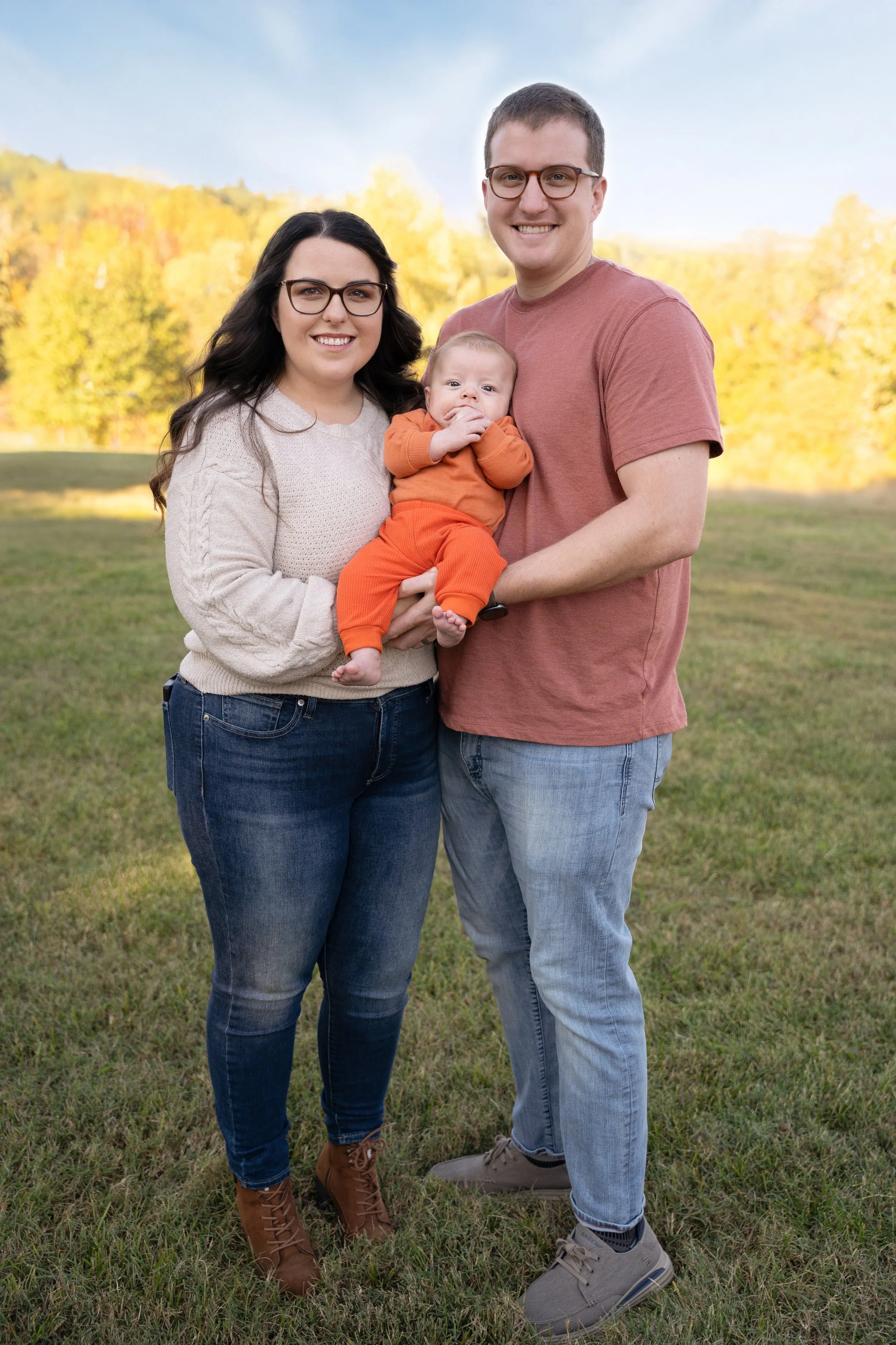 A family of three outdoors in a park on a fall day, with trees in the background with colorful autumn leaves. The woman has long dark hair and glasses, is wearing a cream sweater and jeans. The man has short dark hair and glasses, and is wearing a ru