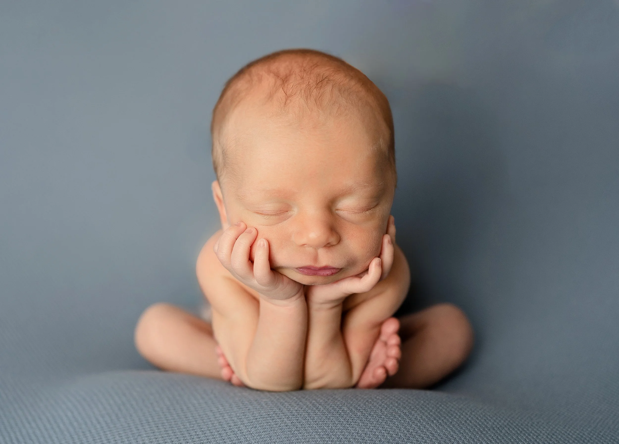 Close-up of a baby with light skin and red hair, resting their face on their hands with eyes closed, against a plain gray background during clayton newborn photographer session