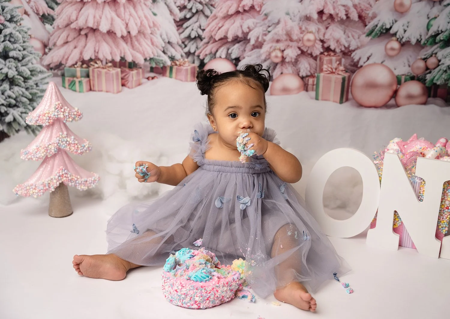 A young girl in a purple dress sitting on a white surface surrounded by Christmas decorations, including a pink glittery tree, wrapped presents, and a large white letter 'O', with a decorated cake and sprinkle-covered sweets nearby.