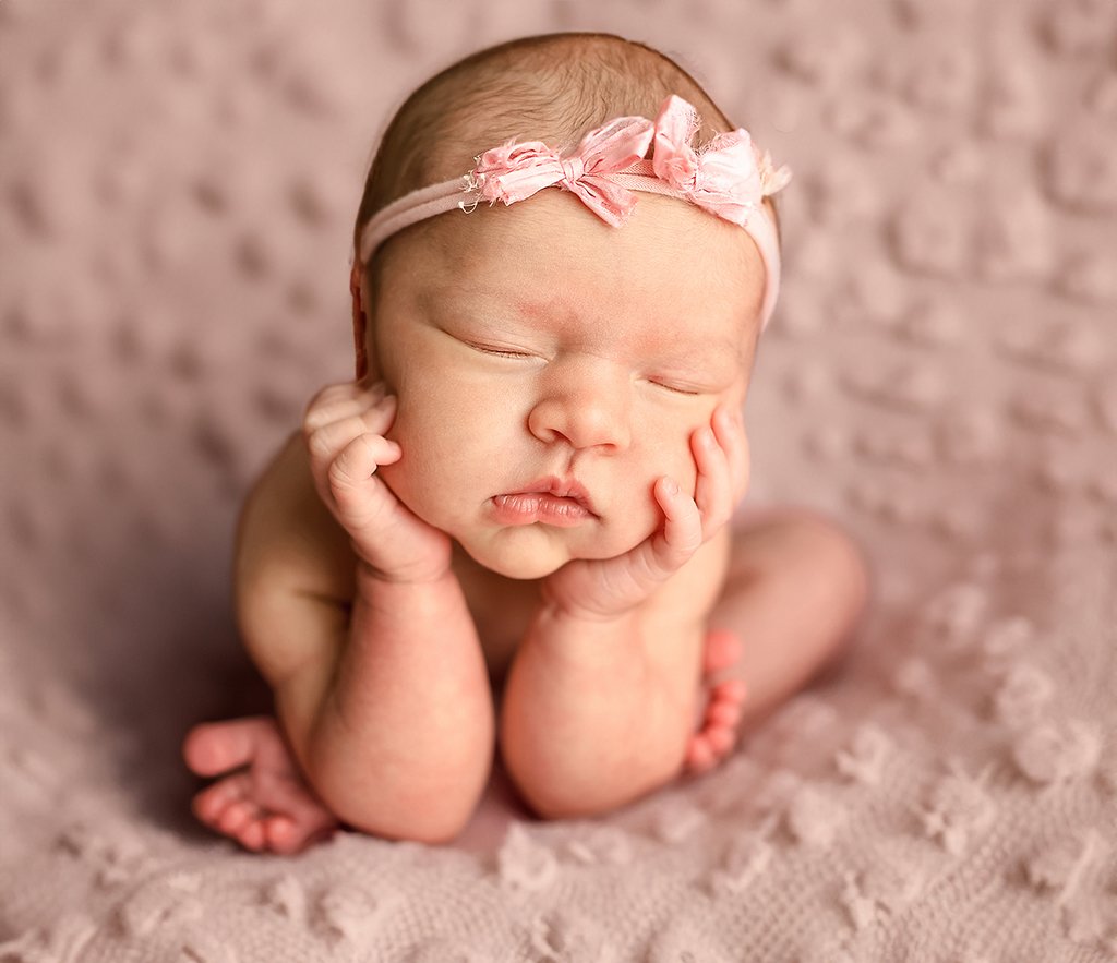 A sleeping newborn baby with a pink headband, resting face on hands, on a pink textured blanket background.