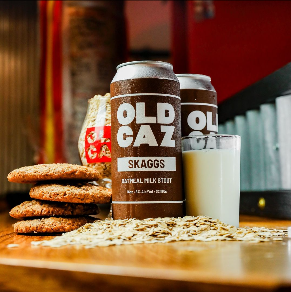 A can of Old Caz Skaggs oatmeal milk stout beer, a glass of milk, and a stack of three cookies on a wooden surface with oat flakes scattered around. In the background, there is a bag of what appears to be rice and a blurred brick wall.