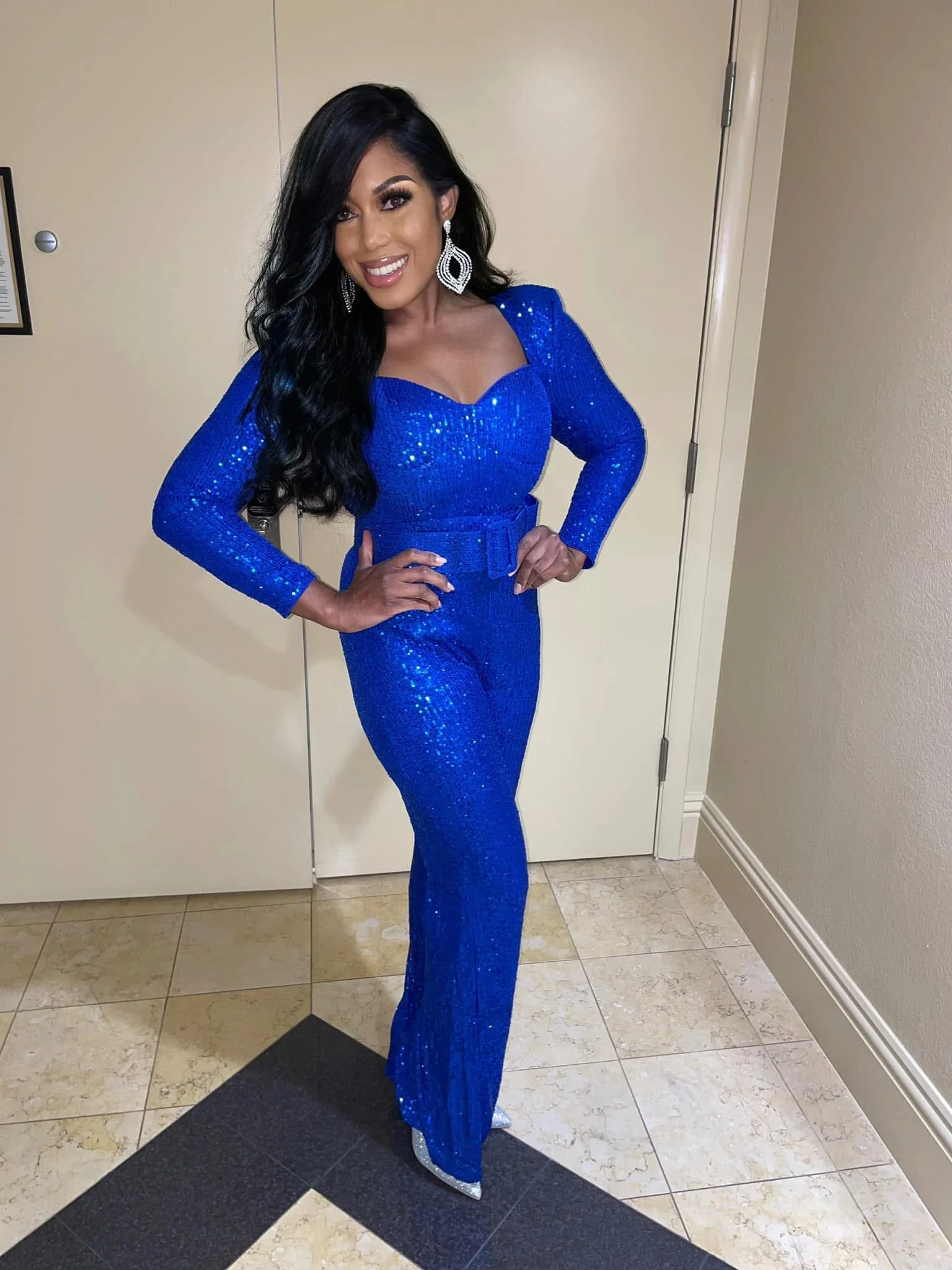 A woman dressed in a shiny, royal blue, sequined jumpsuit with long sleeves, standing indoors near a beige door. She has long, dark wavy hair, large earrings, and is smiling while placing her hands on her hips.