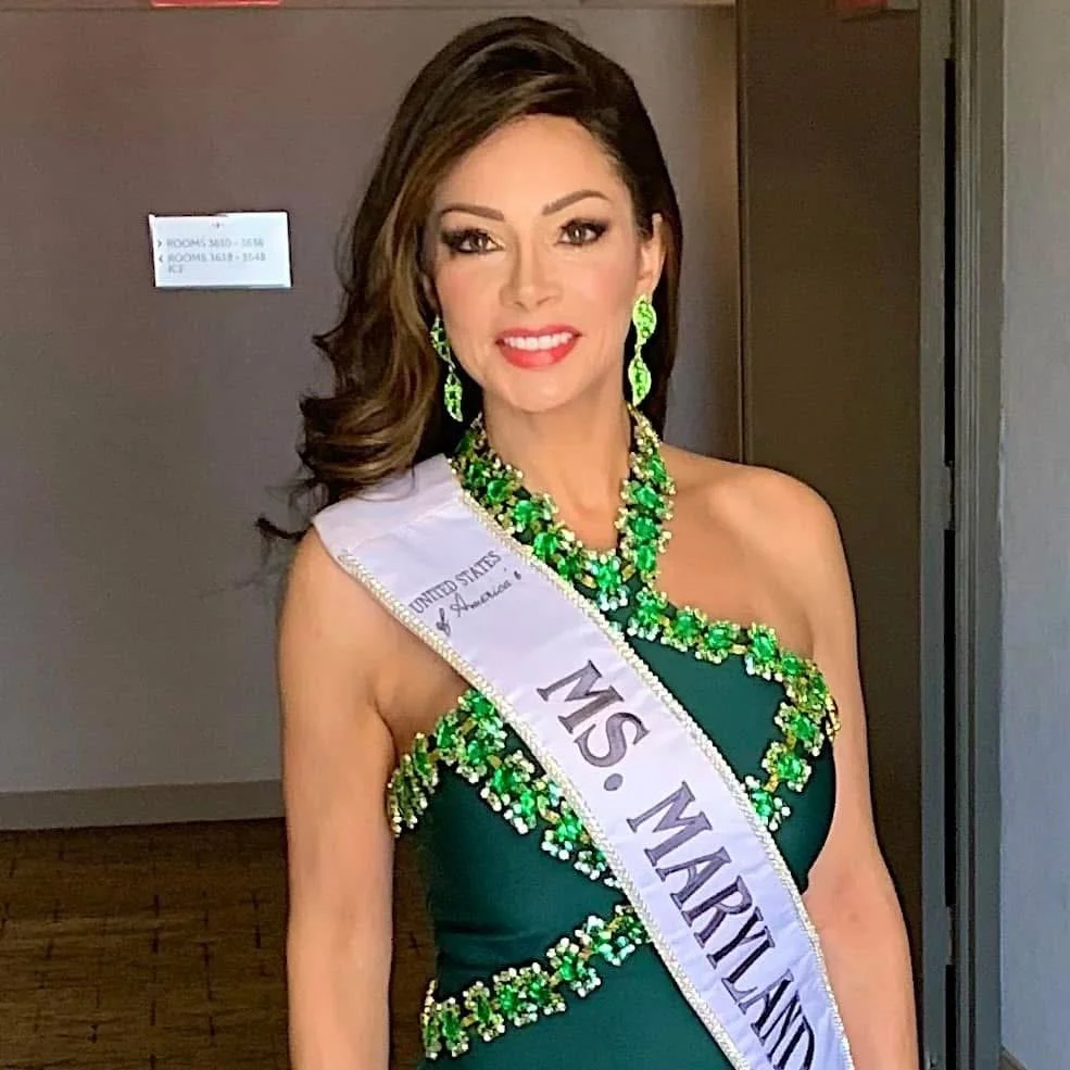 Woman wearing a sash that reads 'Ms. Maryland' and a green dress embellished with green and gold rhinestones, accessorized with matching jewelry, standing indoors with brown walls.
