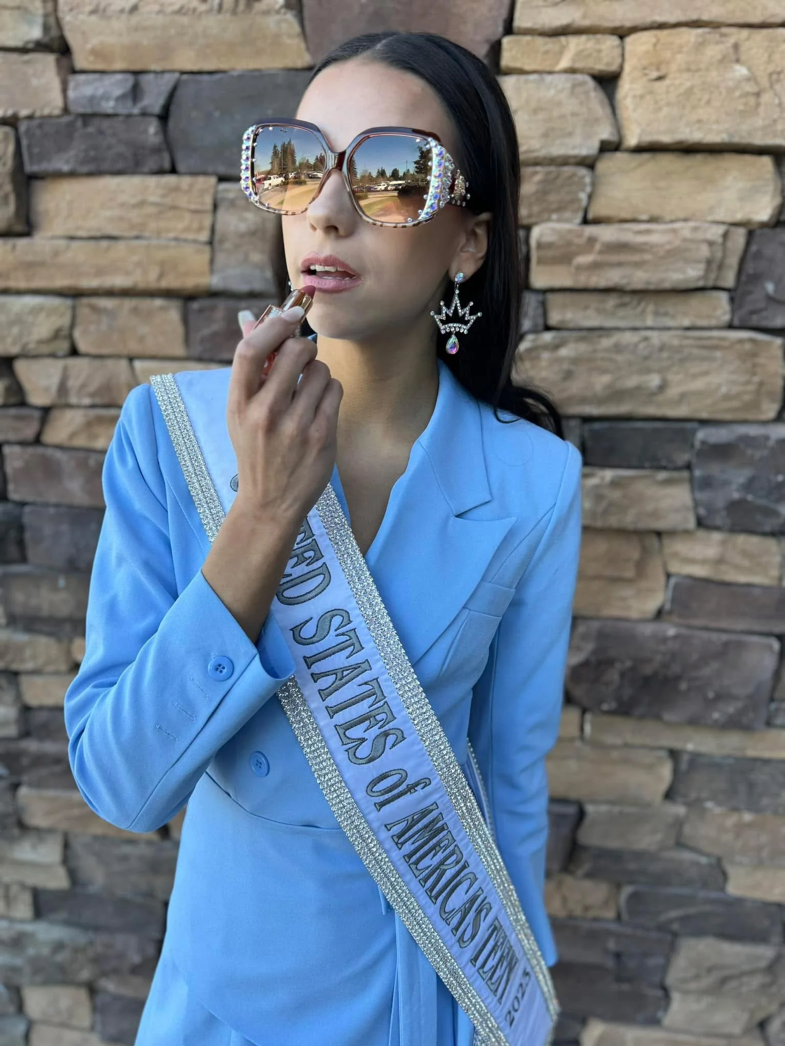 Woman wearing large sunglasses with rhinestones, a blue blazer, and a sash that reads 'Miss World States of America 2023,' standing against a stone wall background, applying lipstick.