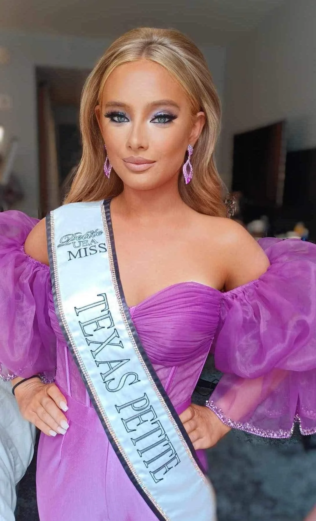 A woman in a purple dress with puffed sleeves, wearing a sash that says 'Miss Texas Petite,' standing in an indoor setting.
