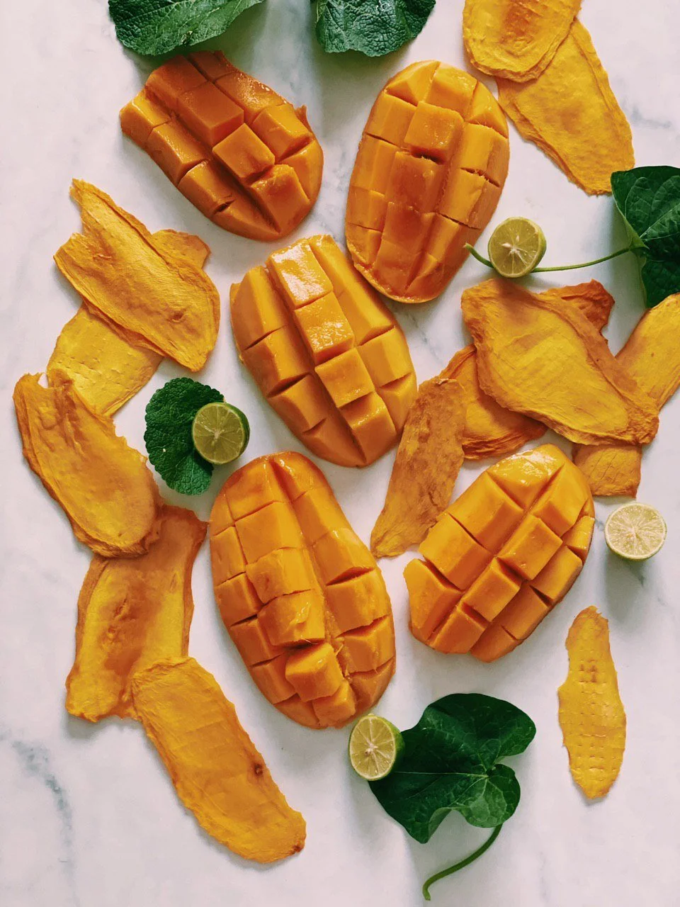 Four halved mangoes with diced flesh on a white surface, garnished with lime slices and green leaves, surrounded by dried mango chips.