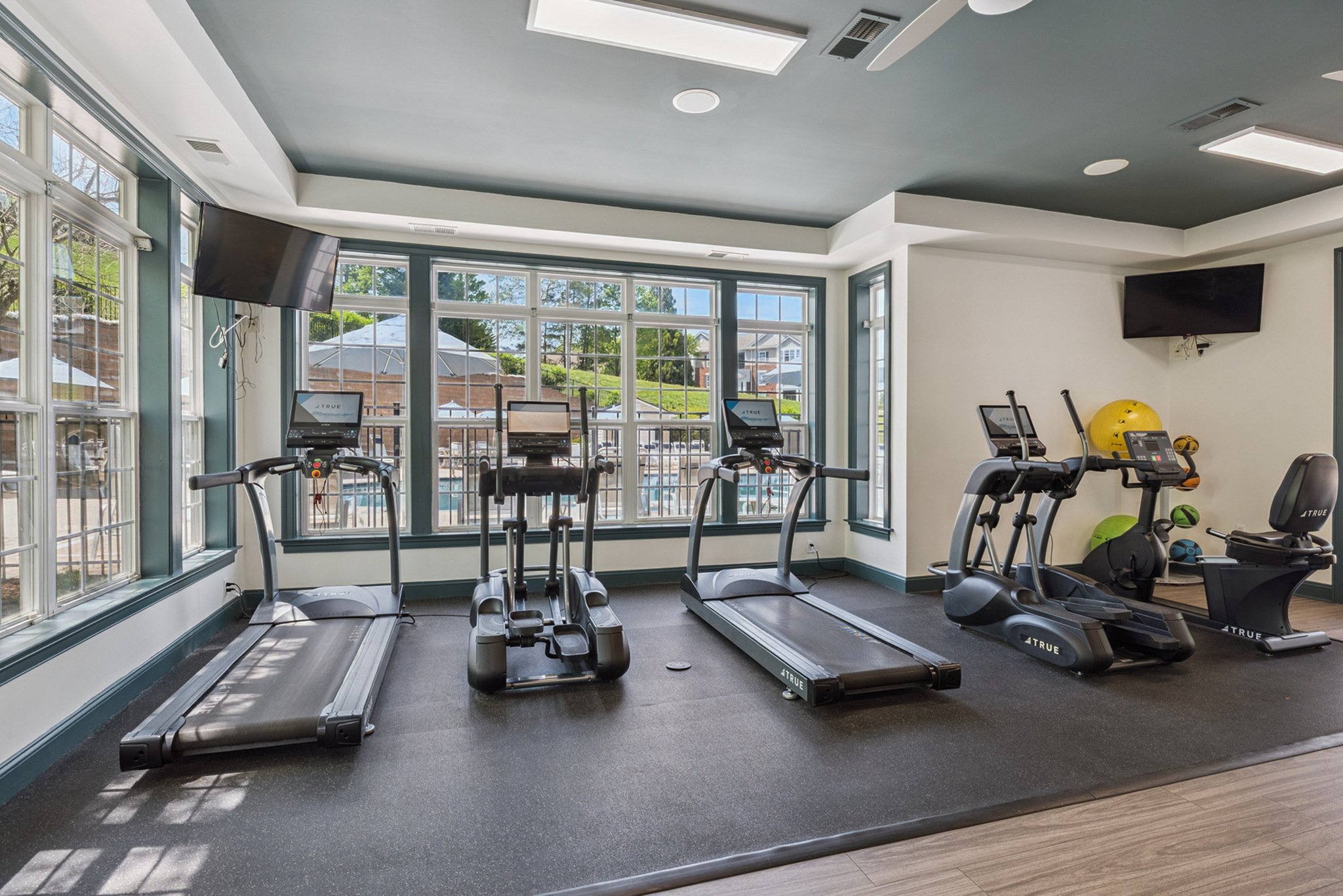 A fitness center with multiple treadmills and exercise equipment in front of large windows with an outdoor swimming pool and patio area visible outside.