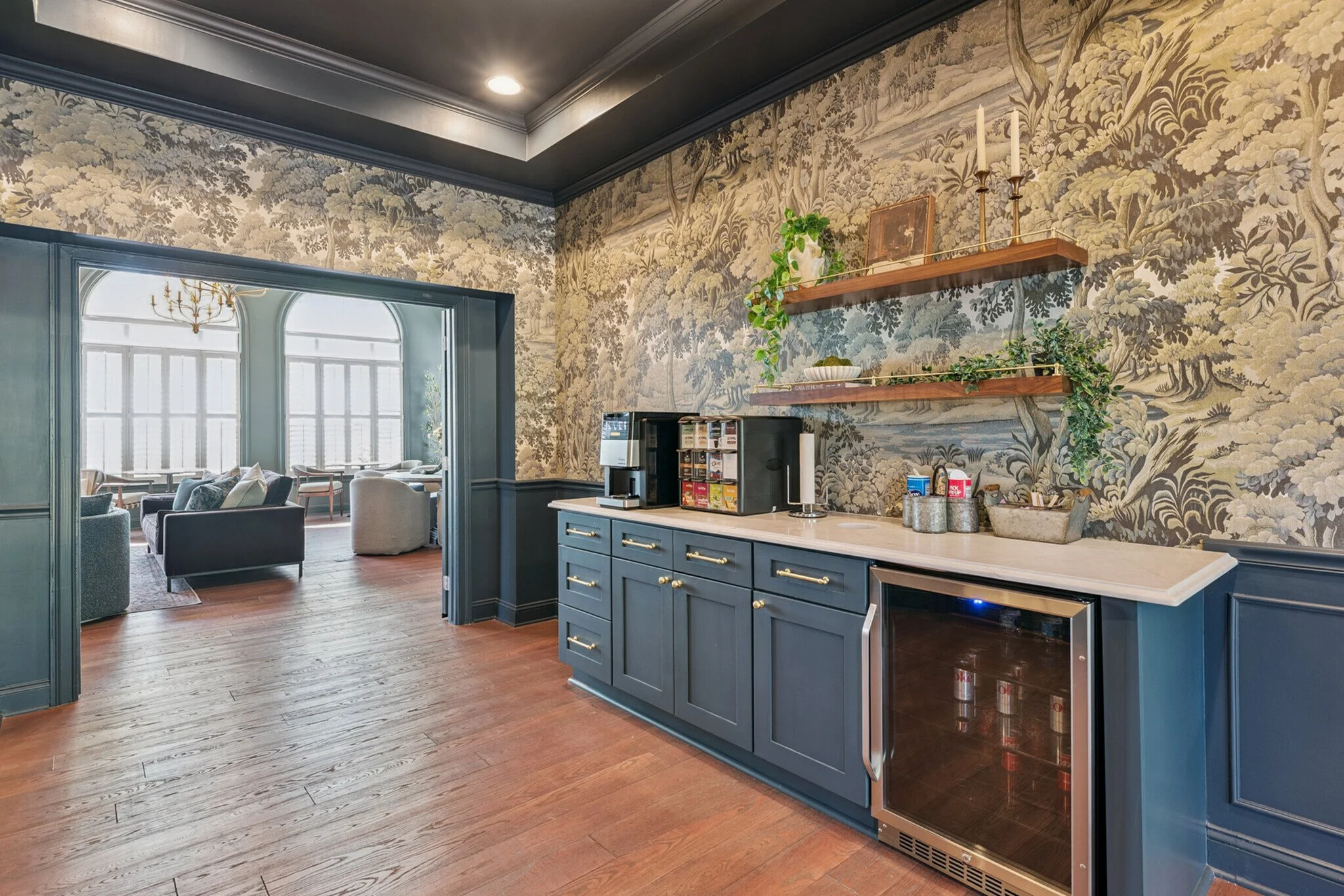 Interior of a room with dark ceiling, wallpaper with a classical landscape pattern, blue cabinetry with gold hardware, a coffee machine, shelves with plants and candle holders, and a view into a bright sitting area with large windows and comfortable 