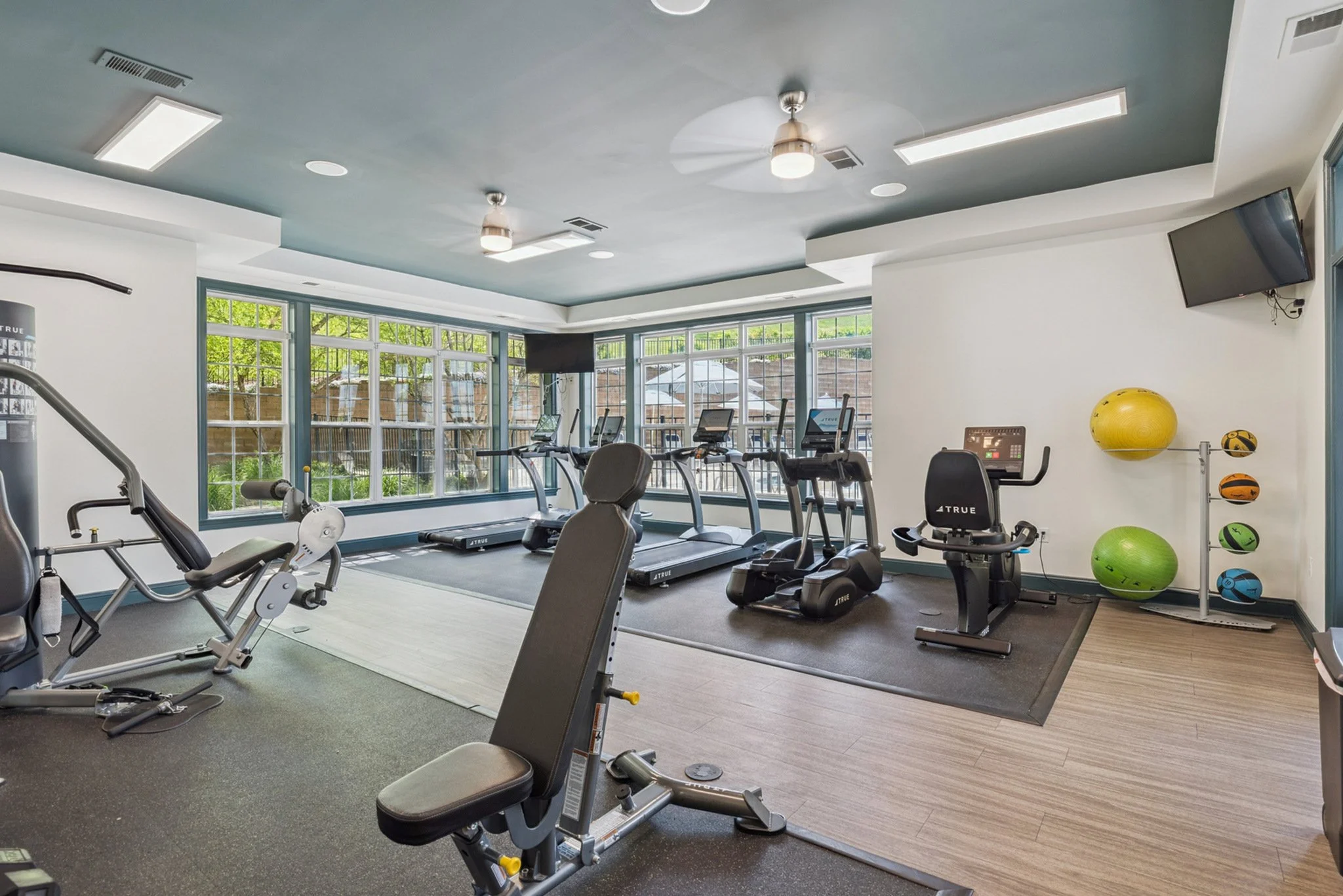 Interior of a fitness room with exercise equipment, including treadmills, elliptical trainers, and stationary bikes. There are large windows with a view outside, and exercise balls and mats are visible on the right side.
