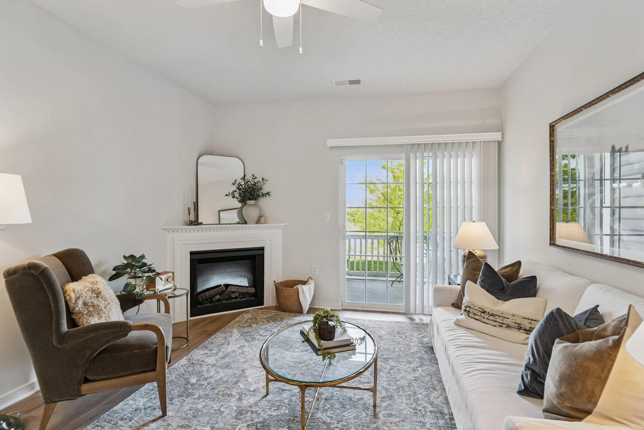 A cozy living room with a beige sofa and dark armchair arranged around a glass coffee table. The room features a white fireplace mantel with a mirror, plants, and decor, and has large sliding glass doors with vertical blinds leading to a balcony with