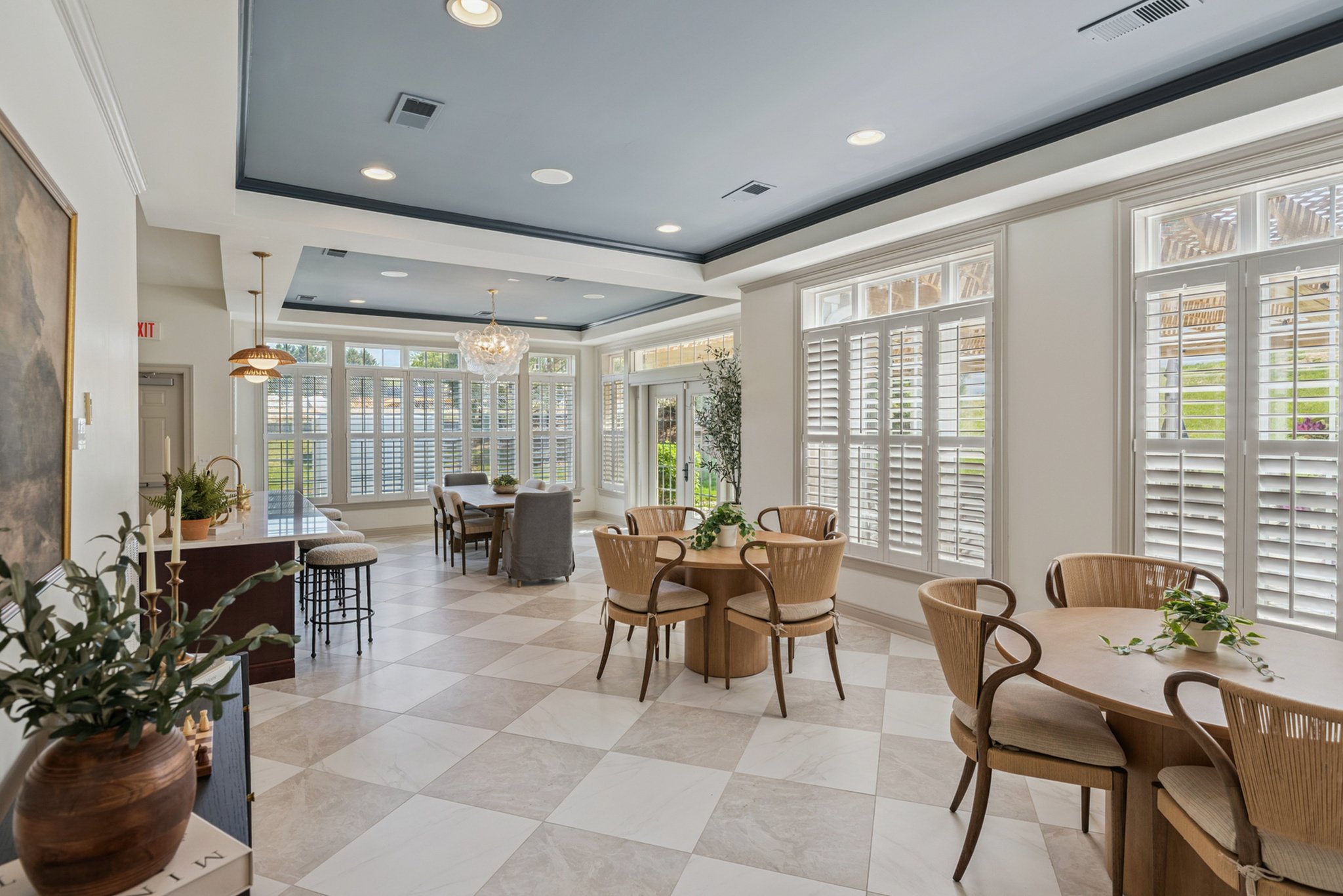 Bright dining area with round and rectangular tables, beige chairs, and large windows with white shutters, decorated with plants and hanging light fixtures.