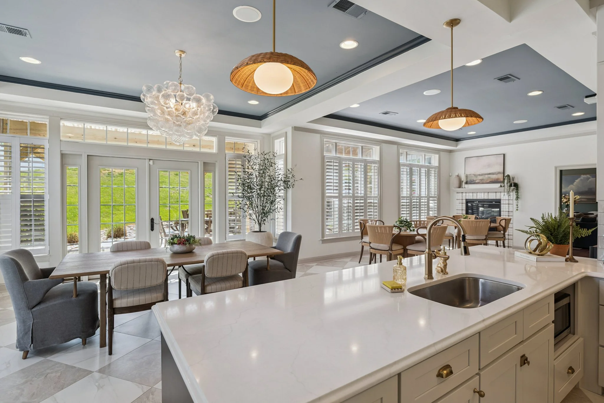 Open-concept kitchen and dining area with white cabinets, a marble countertop, a sink, pendant lights, and large windows with white shutters showing greenery outside.