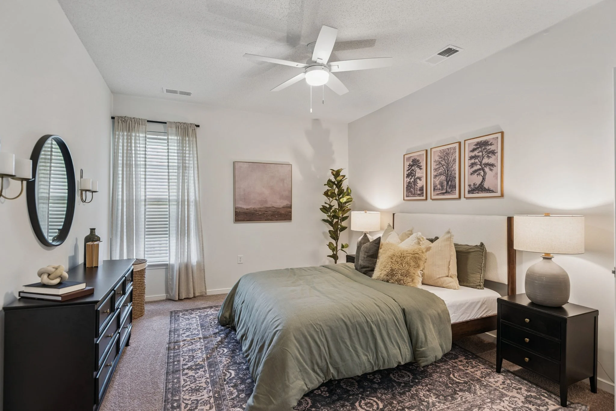 A bedroom with a bed covered in an olive-green duvet, pillows, and a plush throw, flanked by matching black nightstands with lamps, a dresser with decorative items, and a window with sheer curtains and blinds. Artwork and a tall plant decorate the wa