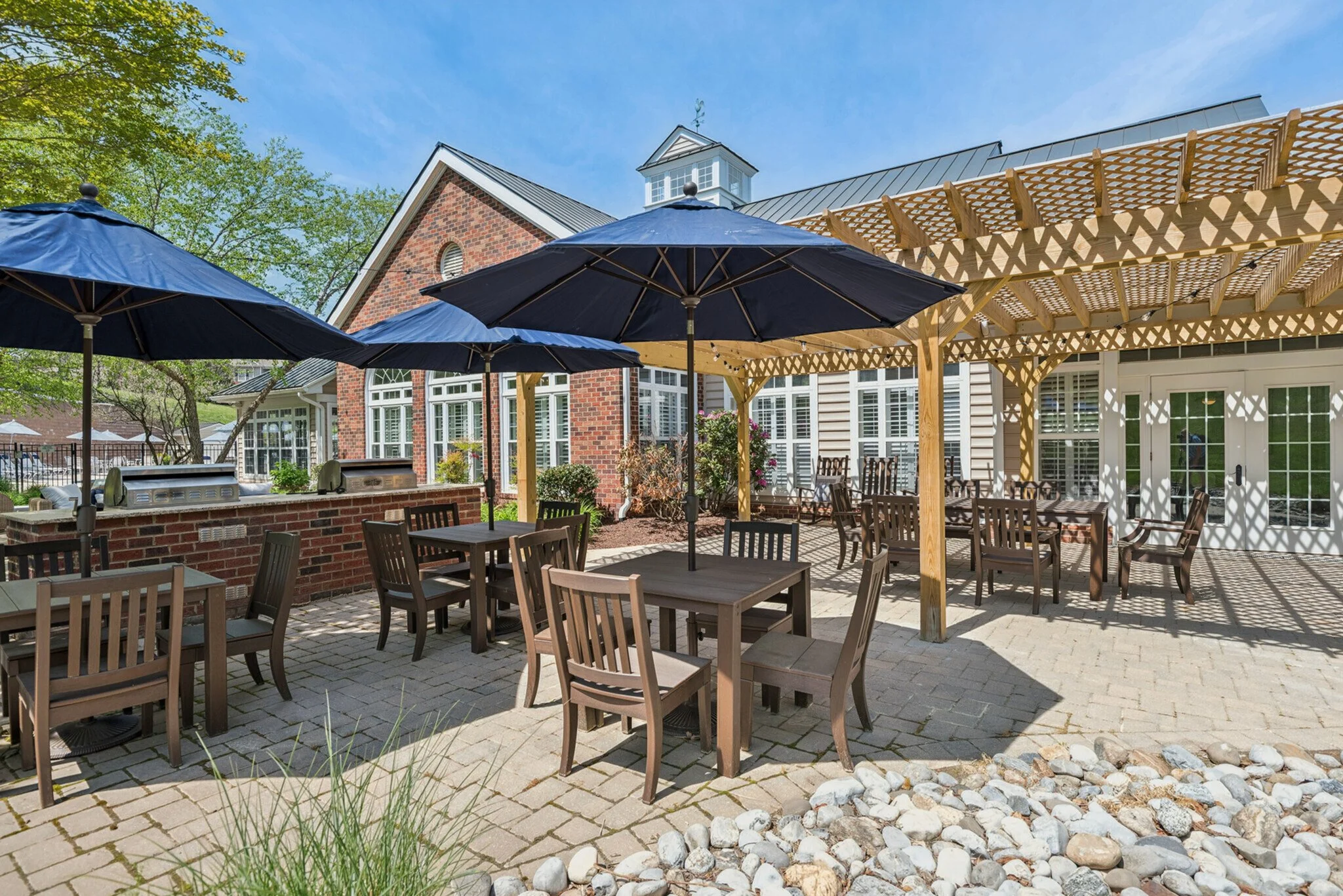 Outdoor patio area with tables, chairs, and blue umbrellas, with a house with brick and siding exterior and a wooden pergola.