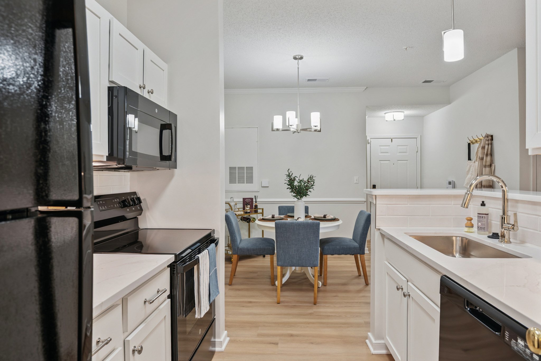 Interior view of a modern kitchen and dining area with white cabinets, black appliances, a white brick backsplash, a sink with a chrome faucet, and a round dining table with blue chairs. The space is well-lit with ceiling lights and a pendant chandel