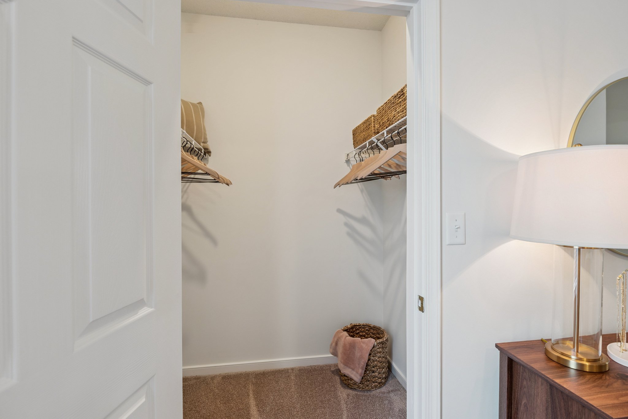 Empty closet with white wire shelves holding baskets and a stack of pillows or linens. A wicker basket with a folded towel is on the floor, and a side table with a lamp is partially visible to the right.