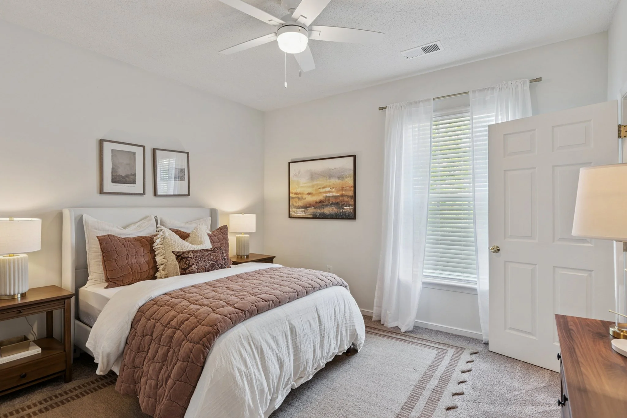 A cozy bedroom with a neatly made bed featuring white and brown bedding, decorative pillows, and two bedside lamps. The room has white walls with framed pictures, a window with white curtains, and a ceiling fan.